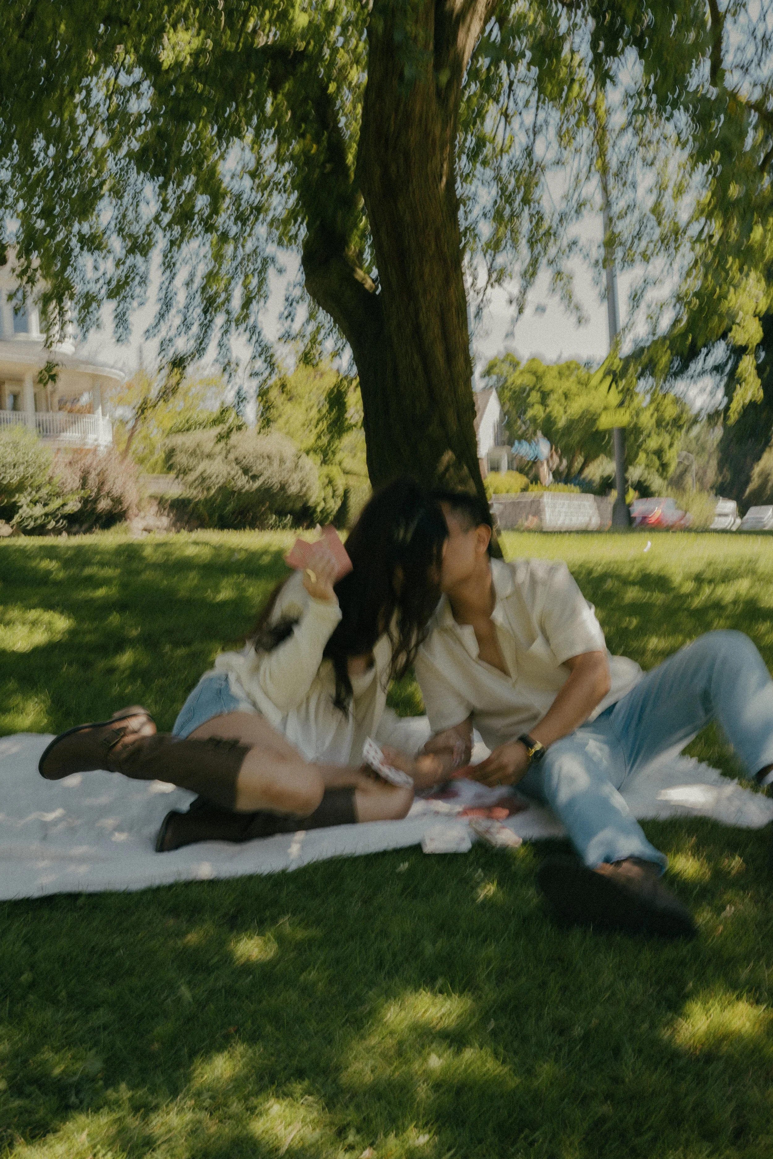Two people sitting on a picnic blanket under a large tree in a park, with green grass and residential houses in the background.