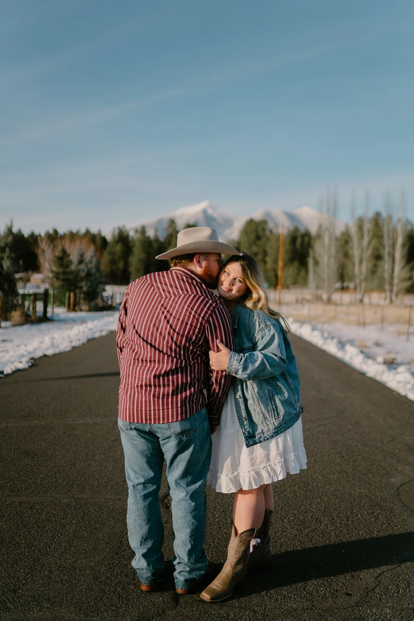 A couple stands close together on a paved road, with snow-capped mountains and a clear blue sky in the background. The man wears a wide-brimmed hat and a red plaid shirt, while the woman wears a denim jacket and a white dress, with cowboy boots.