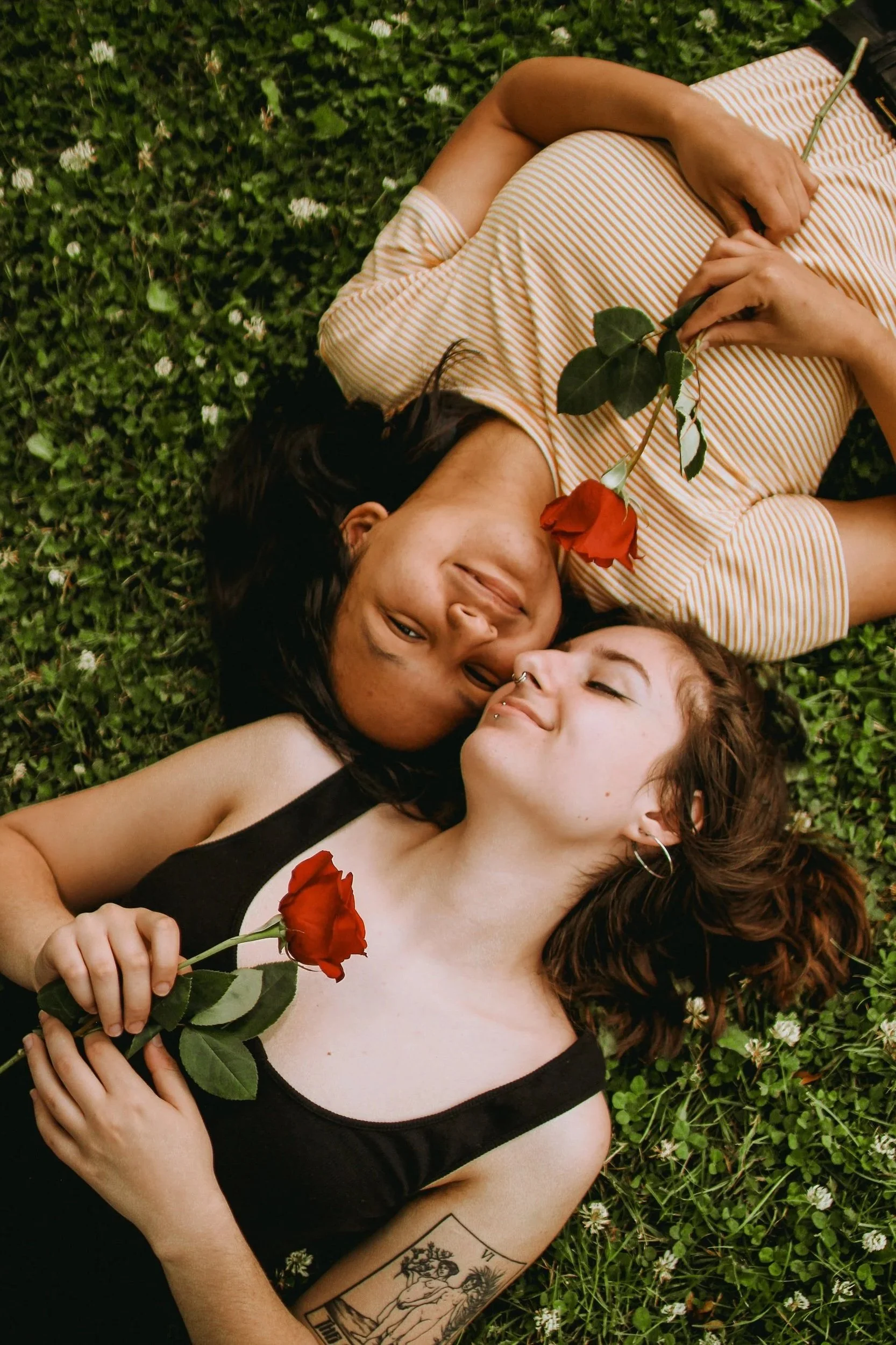 Two women lying on grass surrounded by small white flowers, each holding a red rose, smiling at each other.