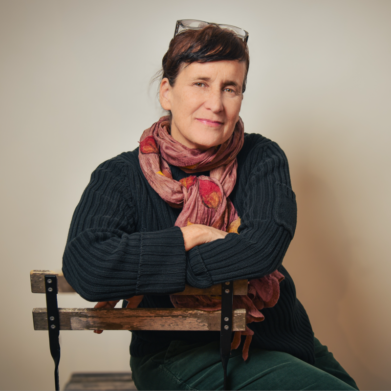 A woman with dark hair, smiling, wearing a denim shirt, against a black background.