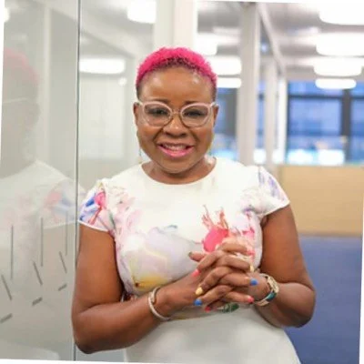 Smiling woman with pink hair, glasses, and floral dress standing indoors with glass wall behind her.
