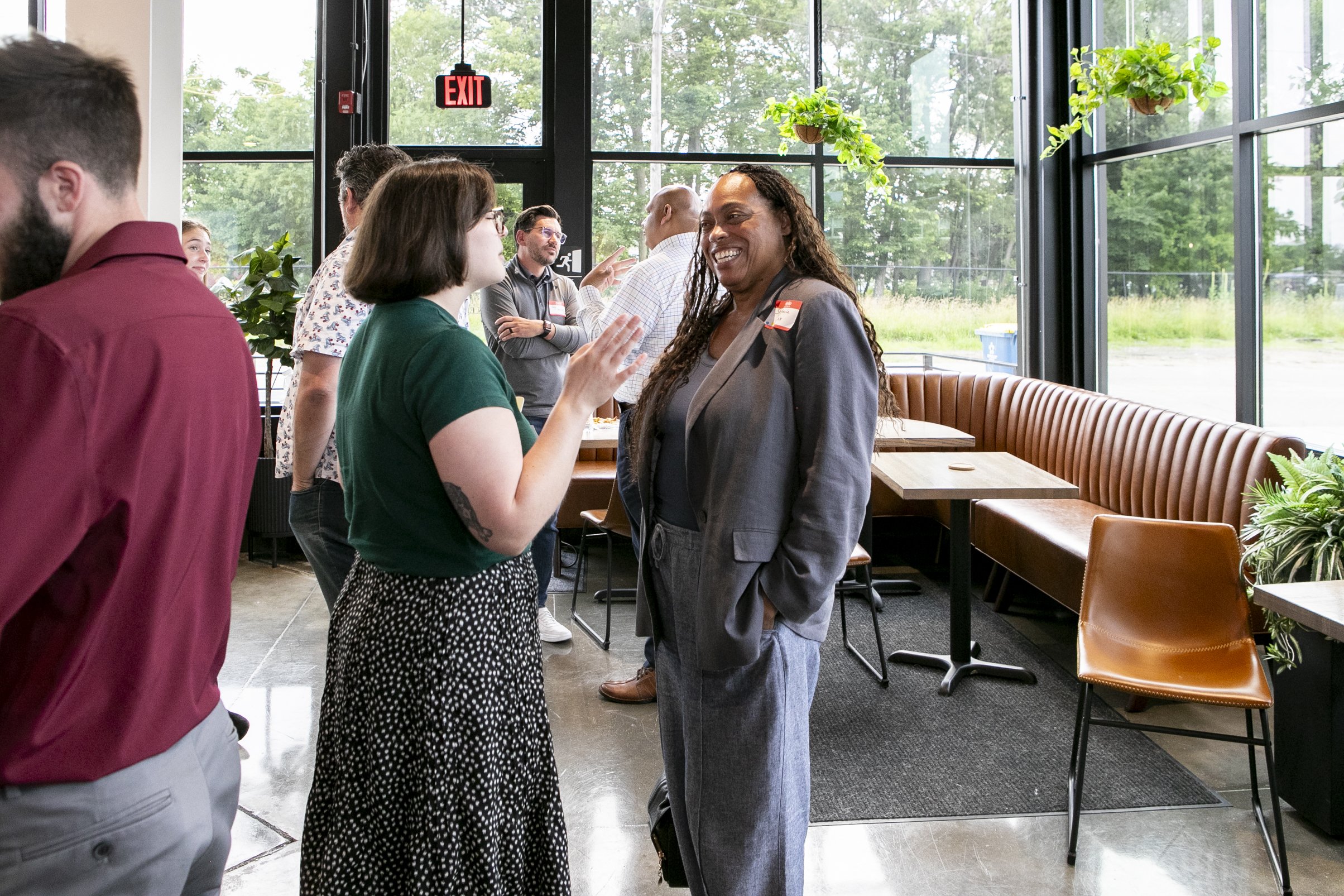 People engaging in conversations inside a modern glass-fronted building with greenery outside, including plants hanging from the ceiling and potted plants.