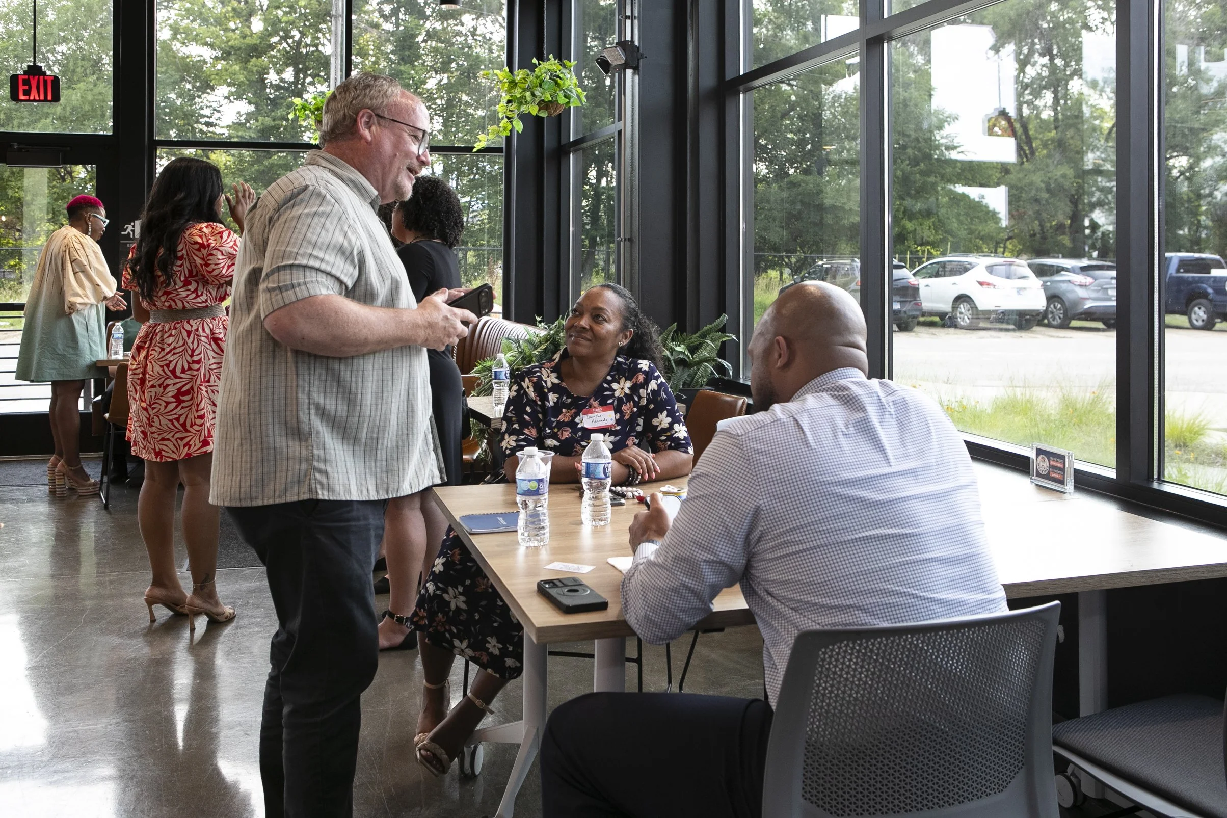 A group of people at a professional event inside a glass-walled building with trees visible outside. Two individuals are seated at a table with water bottles, and a woman is looking at a man standing and engaging with her. Other people are socializing in the background.