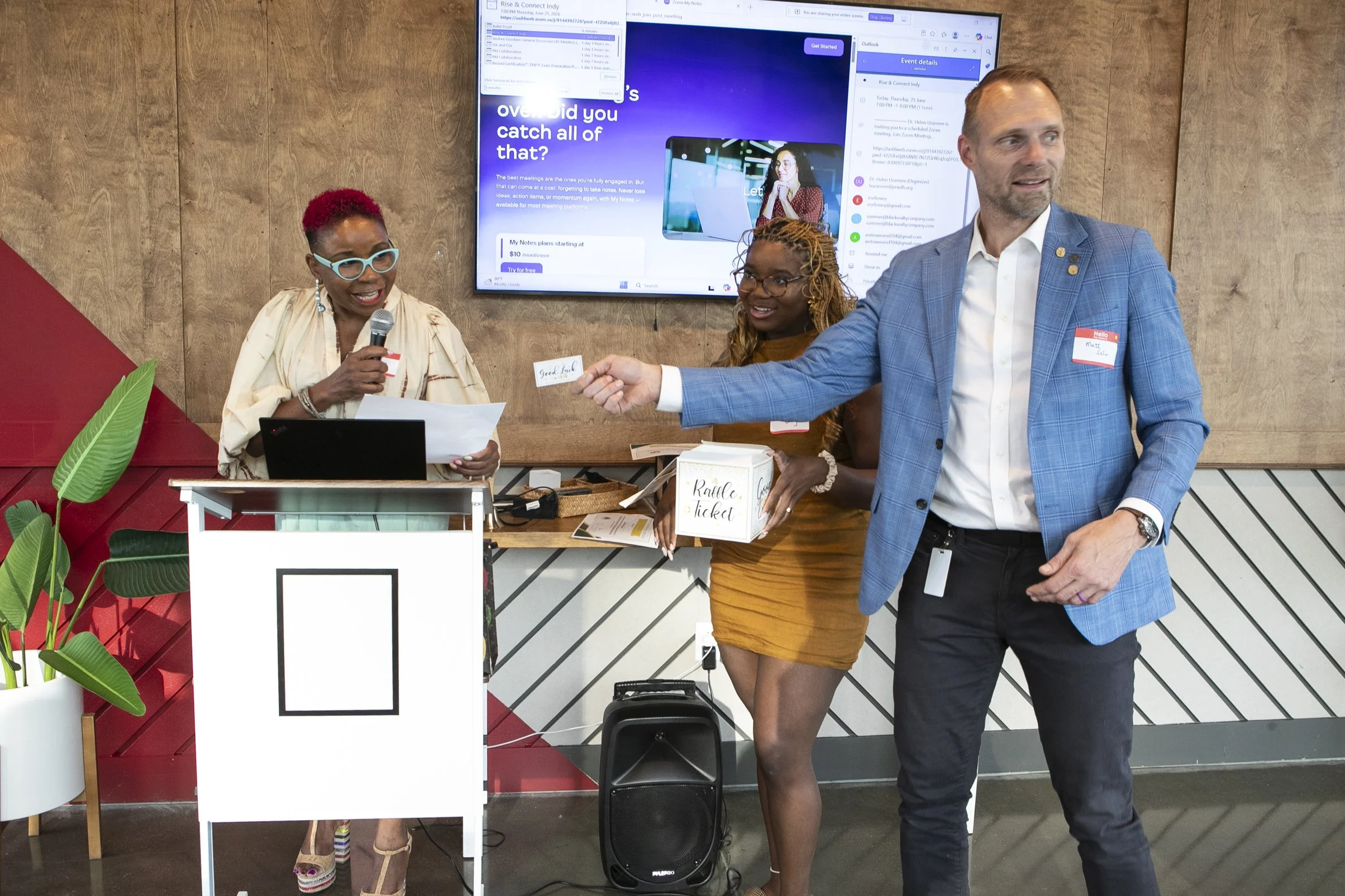 Group of three people at an indoor event; an older woman holding a microphone, a young woman in a mustard dress holding a raffle box, and a man in a blue blazer extending a hand; a large screen behind them displays a presentation and event details.