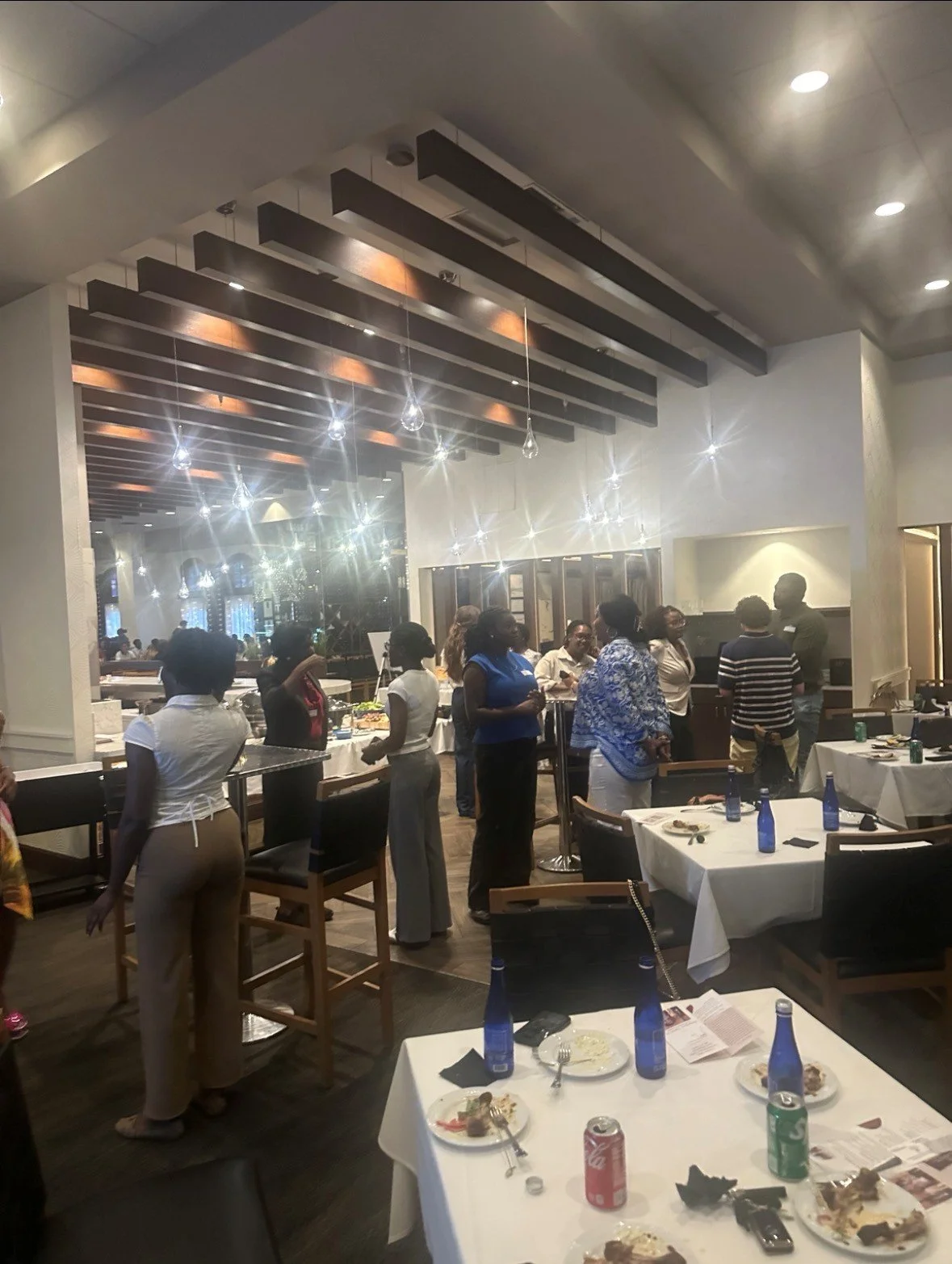 People gathered in a restaurant or banquet hall, socializing and standing around tables with plates and drinks, some plates with leftover food, in a well-lit space with modern decor.