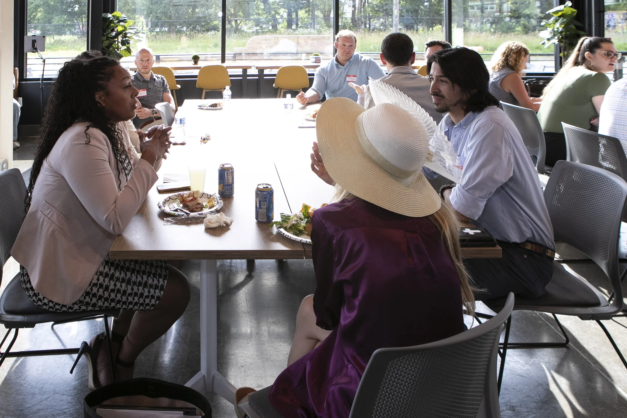 People sitting around a conference table engaged in discussion, with plates of food and drinks in front of them, inside a room with large windows and yellow chairs.