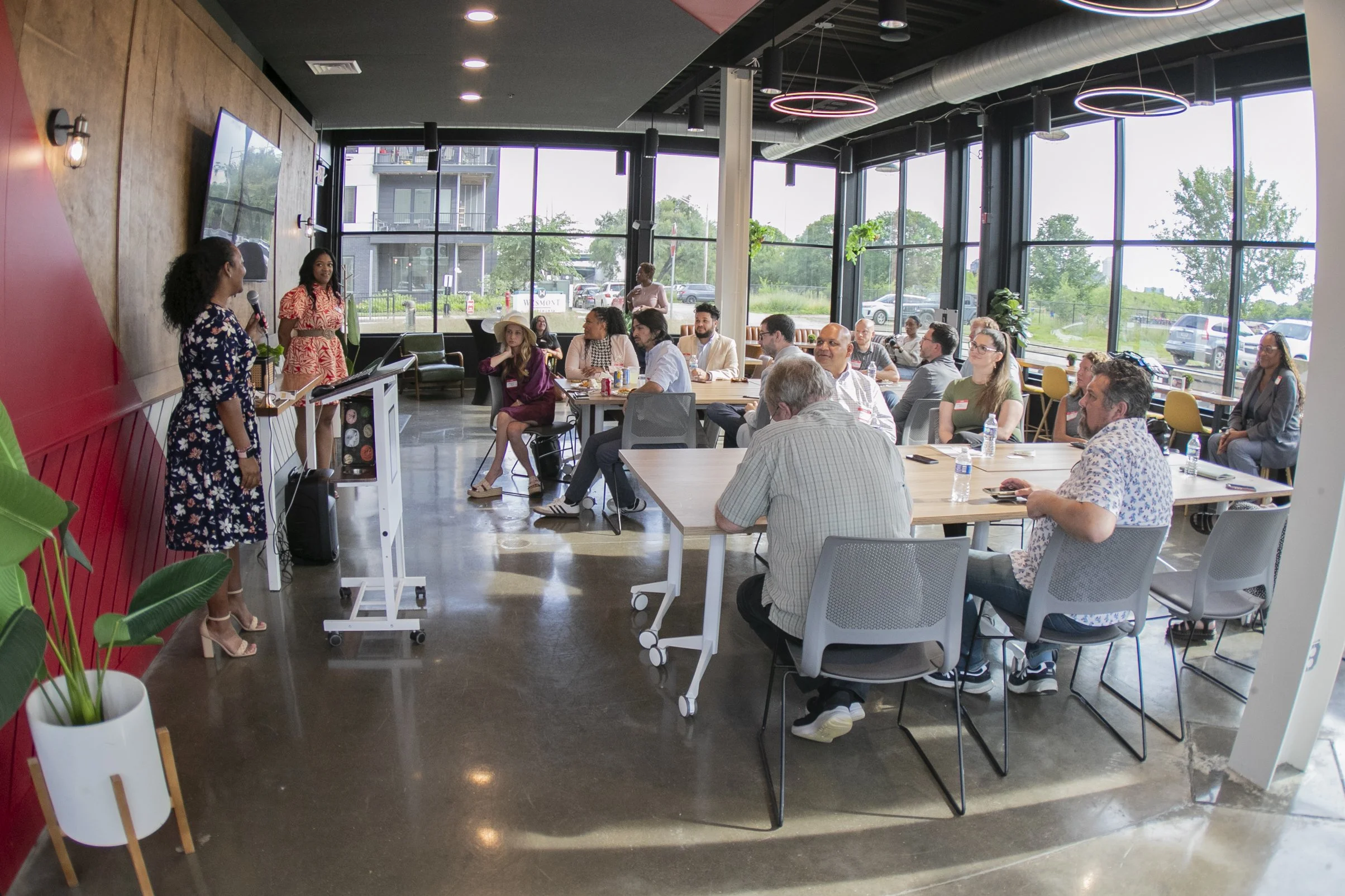 A diverse group of people attending a presentation or meeting in a modern, brightly lit conference room with large windows, indoor plants, and contemporary lighting fixtures.