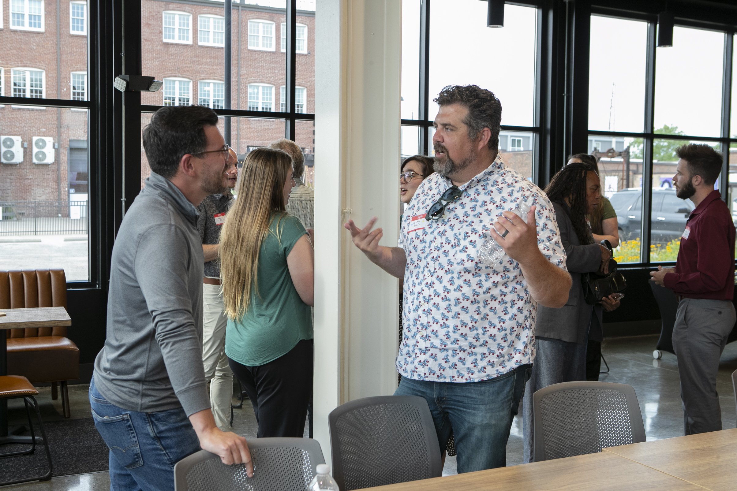 People engaging in conversation at a networking event or workshop inside a modern building with large windows showing a cityscape outside.