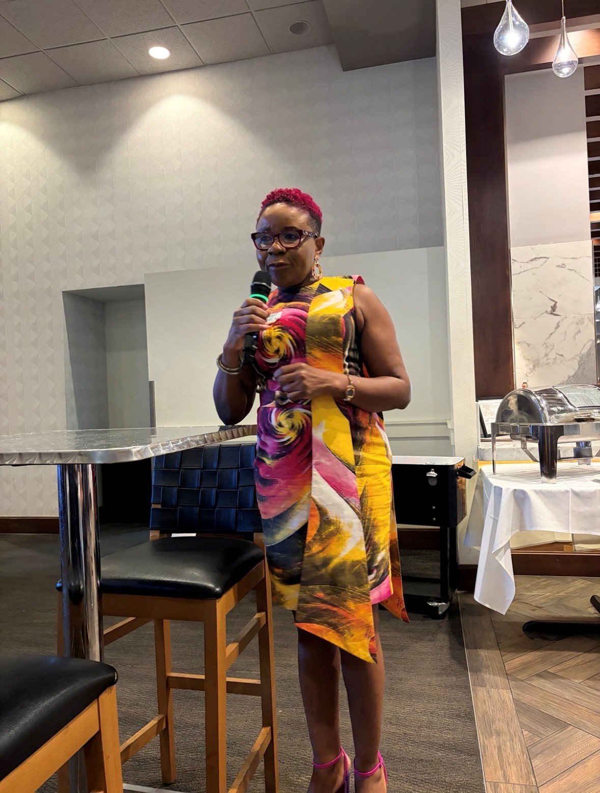 A woman with short pink hair, glasses, and earrings, holding a microphone in a restaurant or event space. She is wearing a colorful, abstract-patterned dress with pink, yellow, and black tones, and pink high-heeled shoes. There are chairs, tables, and chafing dishes in the background.