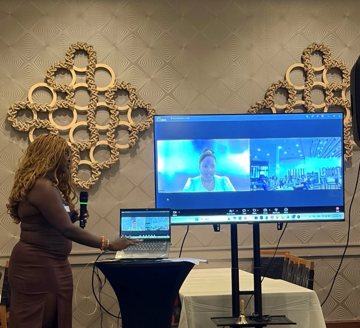 Woman with long, curly hair standing with a microphone, presenting a virtual meeting or conference with screens showing multiple participants, in a decorated room with geometric wall art.