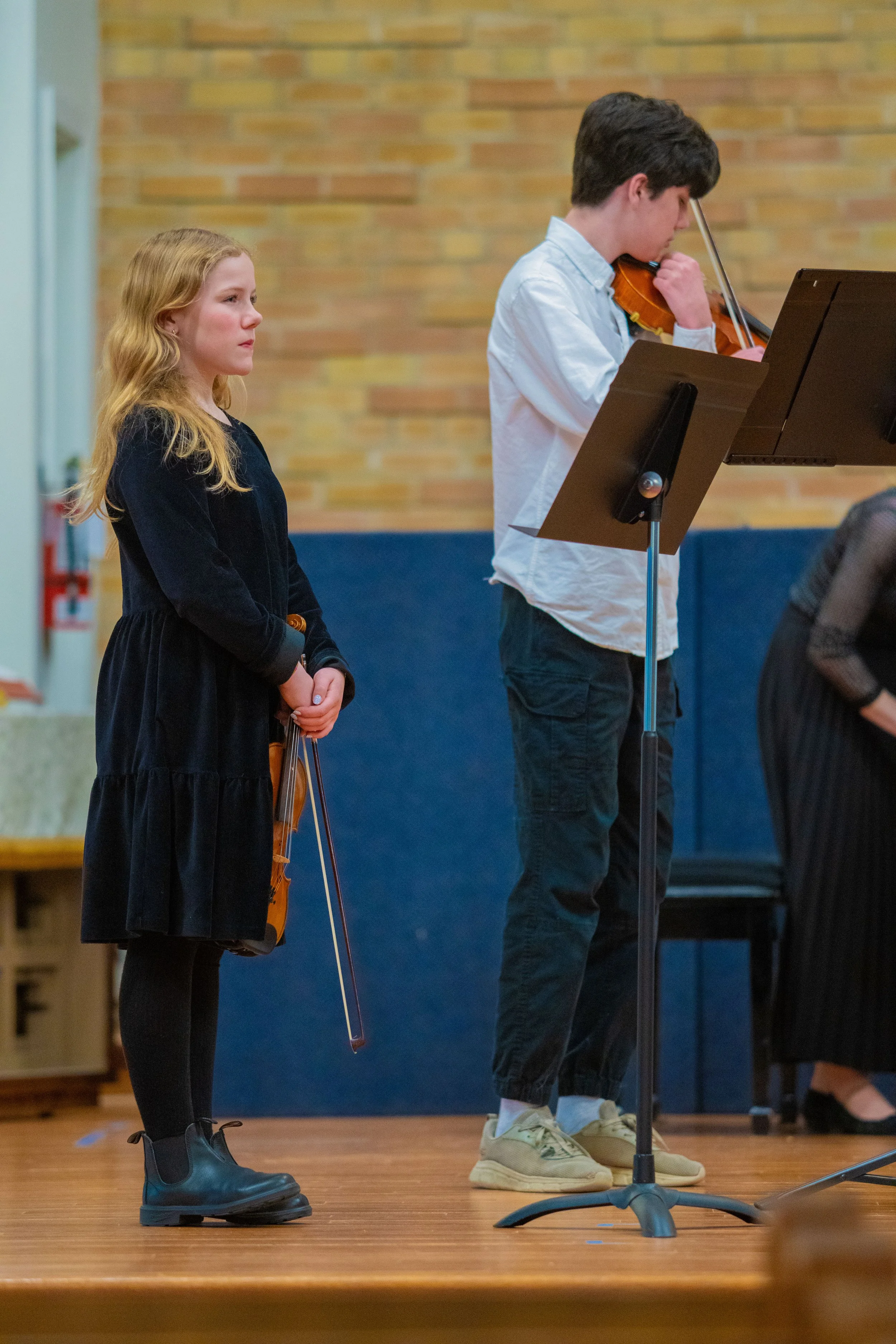 A young girl looks with peaceful focus while a teenage boy tunes his violin before a concert