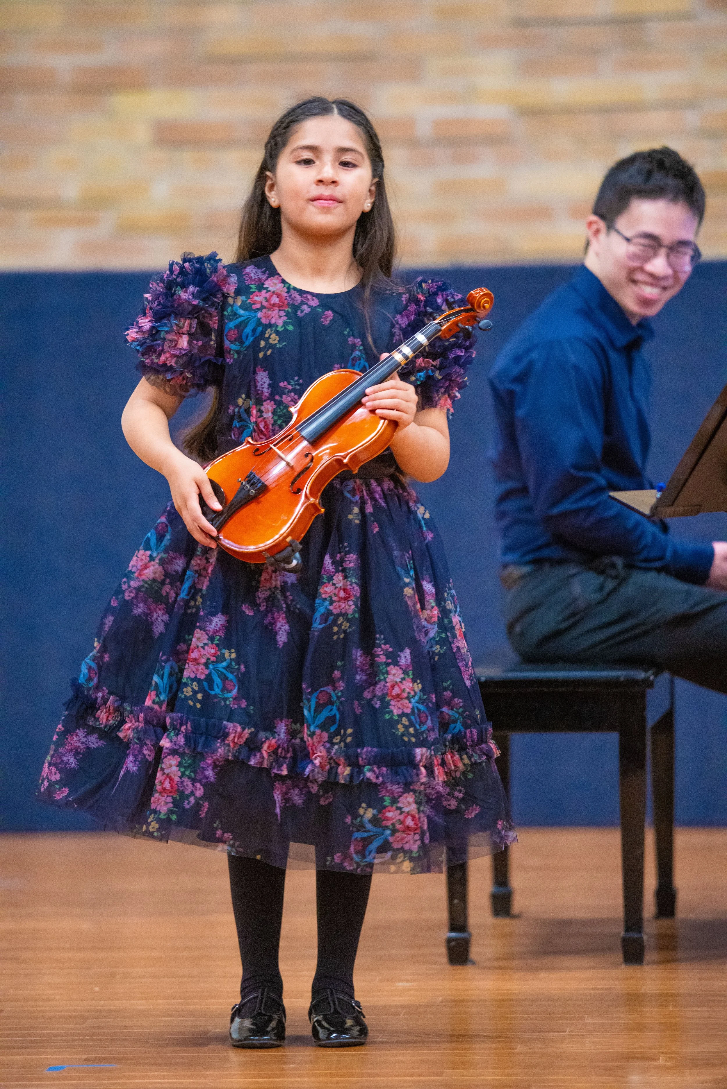 A young child looks proudly after performing at a violin recital while a piano accompanist smiles on