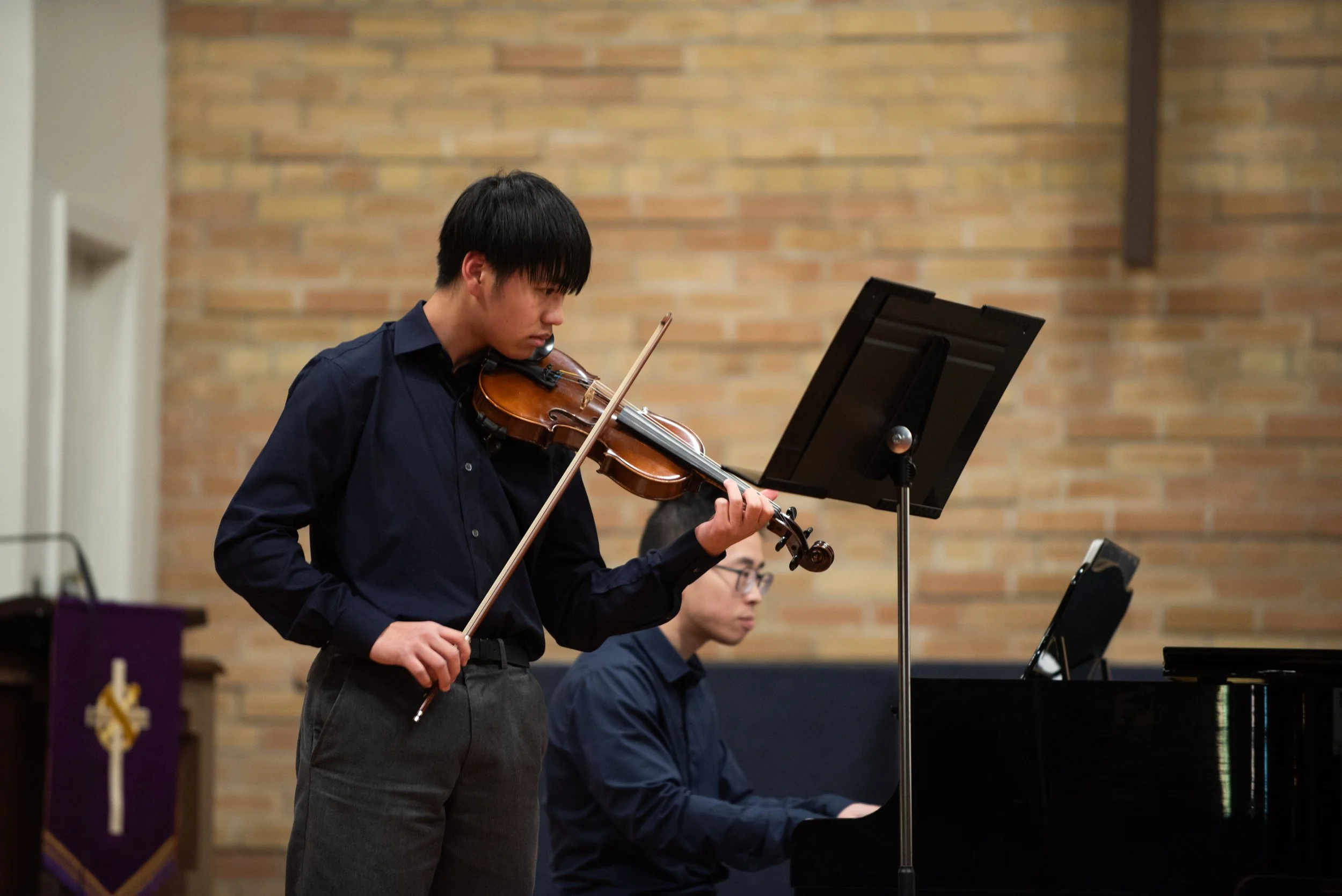 A teenage boy plays a violin solo with focus on stage at a recital along with a piano accompanist