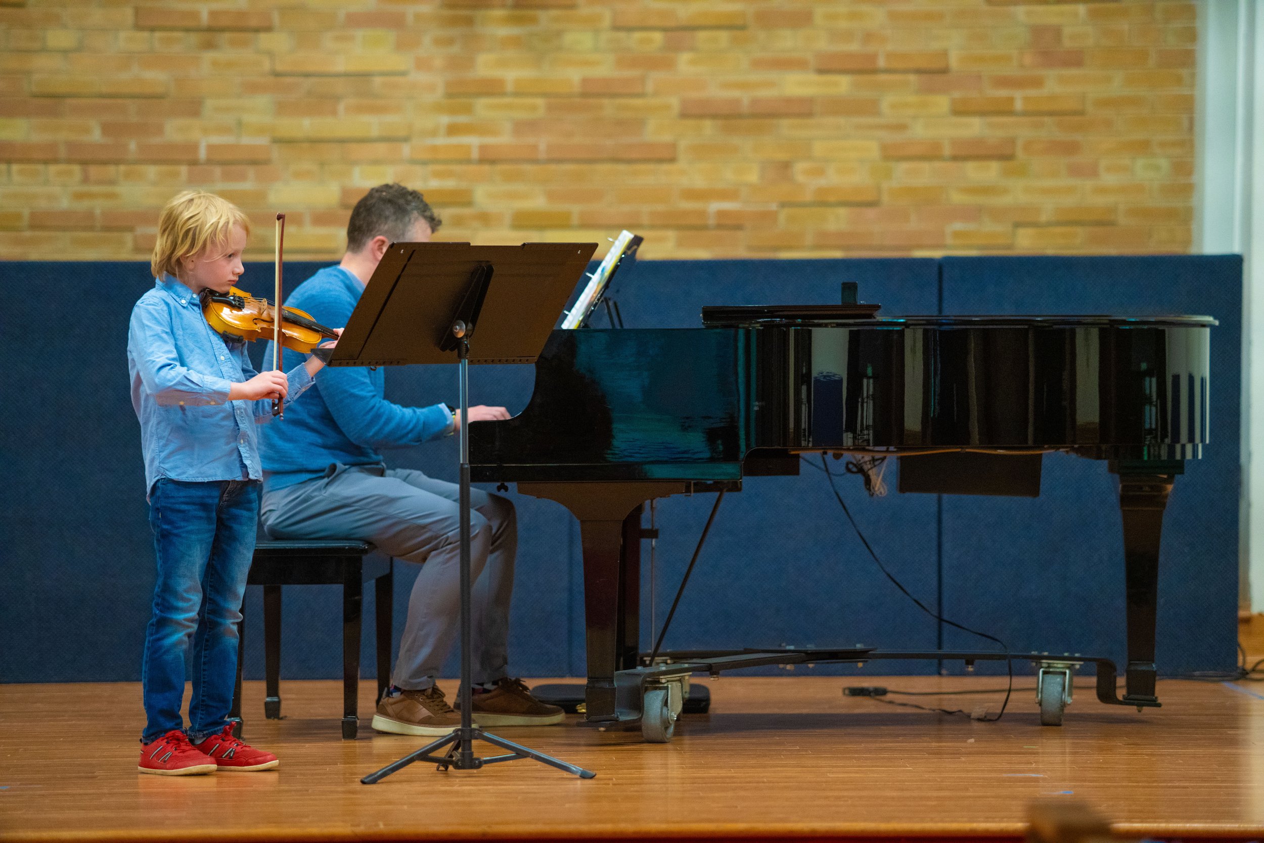 A young boy plays violin on stage with his father playing piano at a recital performance.