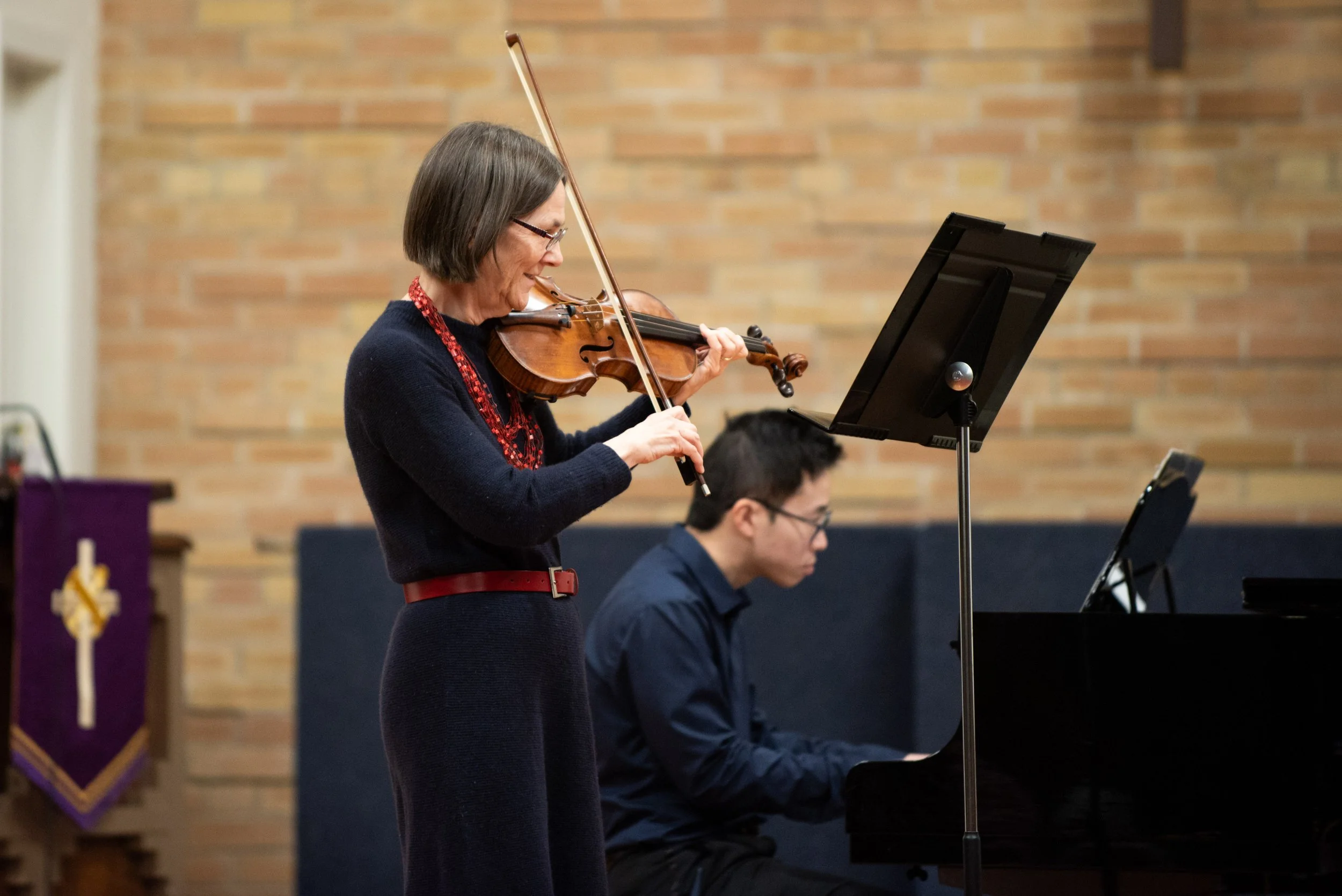 An adult student plays violin on stage in a recital while smiling with a piano accompanist