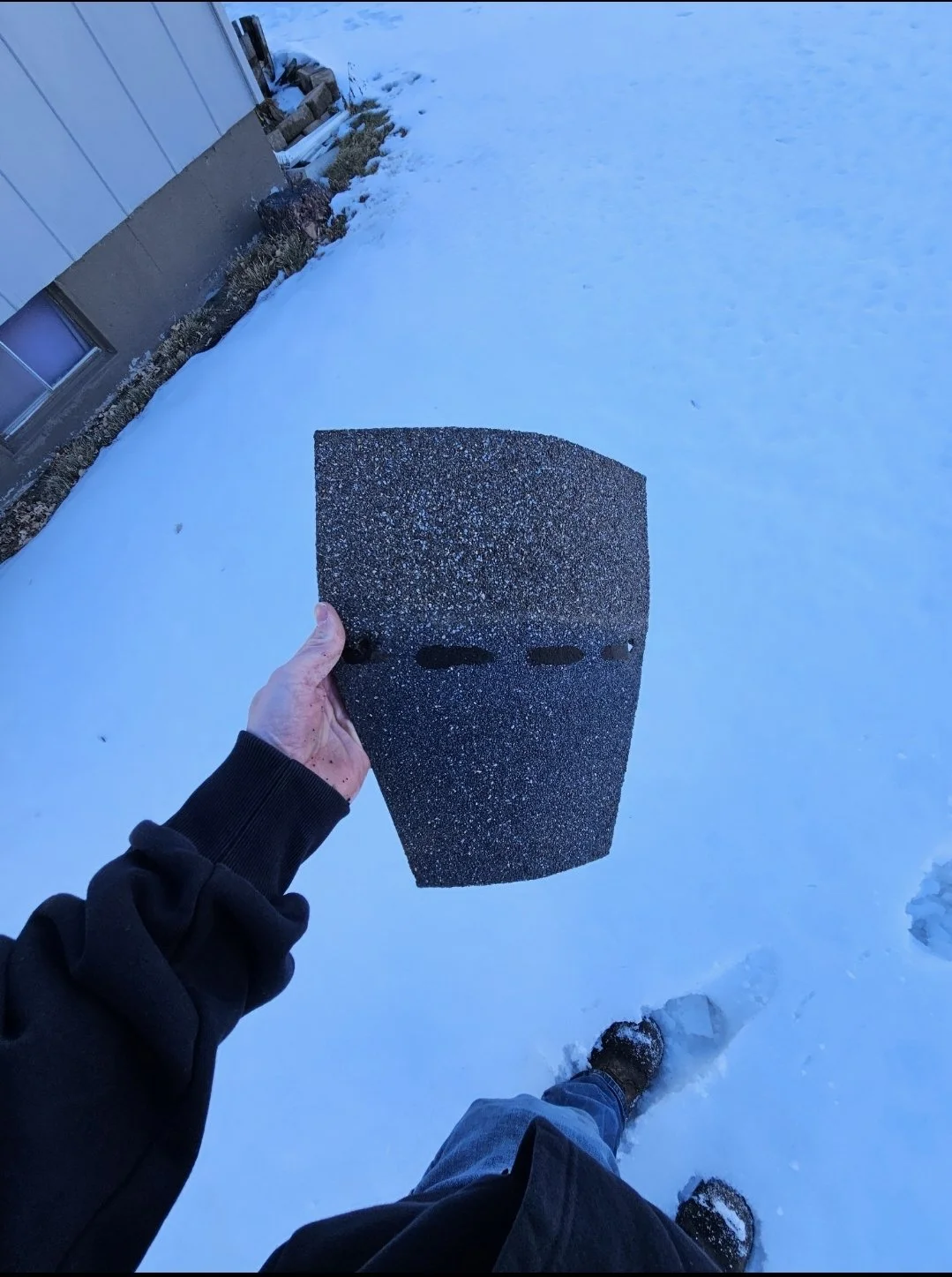 Person holding a large piece of roofing shingle outdoors on snow-covered ground near a building.