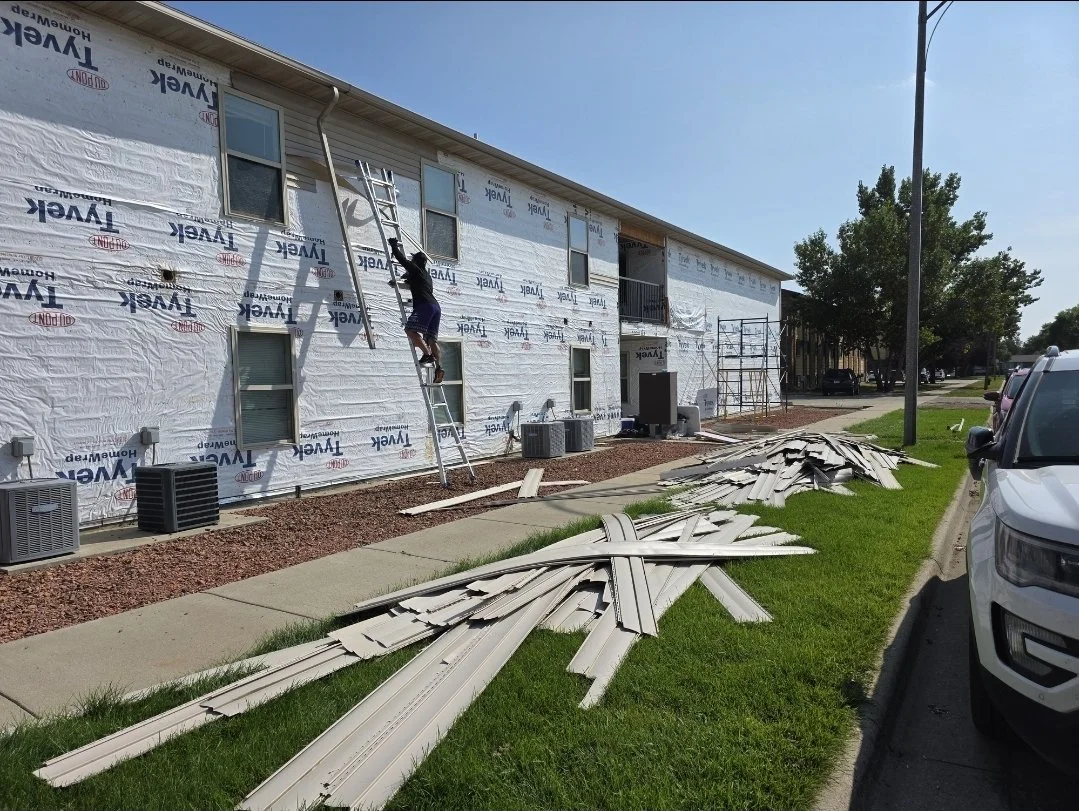 A person standing on a ladder installing or repairing the exterior of a building that is covered with a weather-resistant house wrap. There are several pieces of siding or trim on the ground nearby, and construction scaffolding is set up along the side of the building. Parked cars and a grassy area are also visible.