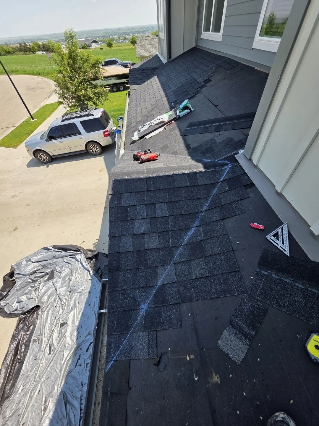 View from a rooftop showing roofing repair work, tools, and materials with a driveway, parked cars, grassy area, and houses in the background.