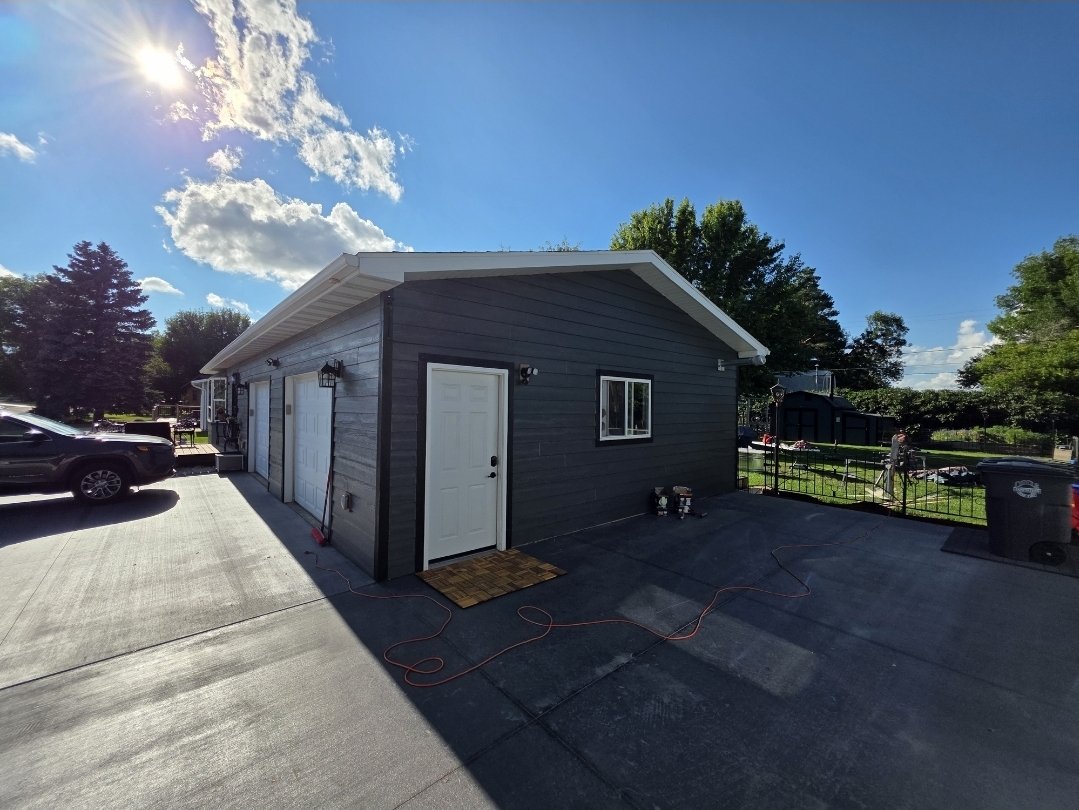 A small gray building with white doors and a window, surrounded by a driveway and some parked cars, under a sunny sky with scattered clouds.