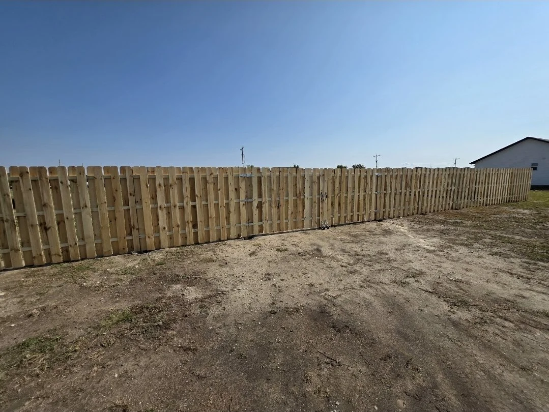 A newly built wooden fence in an open yard with a clear blue sky and a neighboring house in the background.