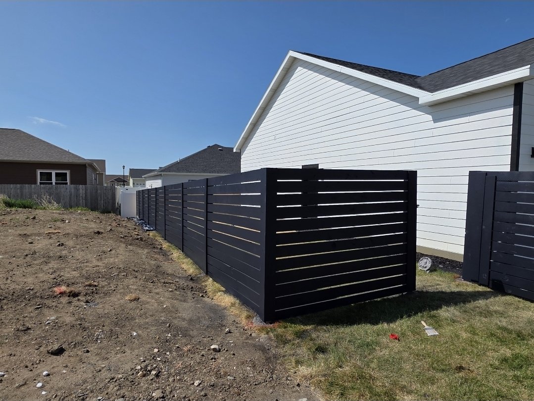 A black privacy fence beside a white house with siding, on a partly cloudy day. The ground on one side of the fence is dirt, and on the other side, there is grass.