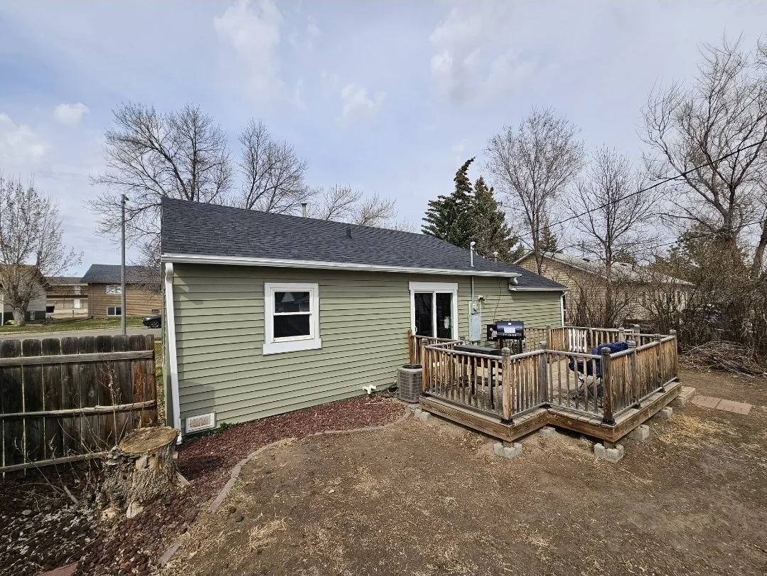 Backyard view of a house with green siding, a small wooden deck with patio furniture, a grill, leafless trees, and neighboring houses in the background.