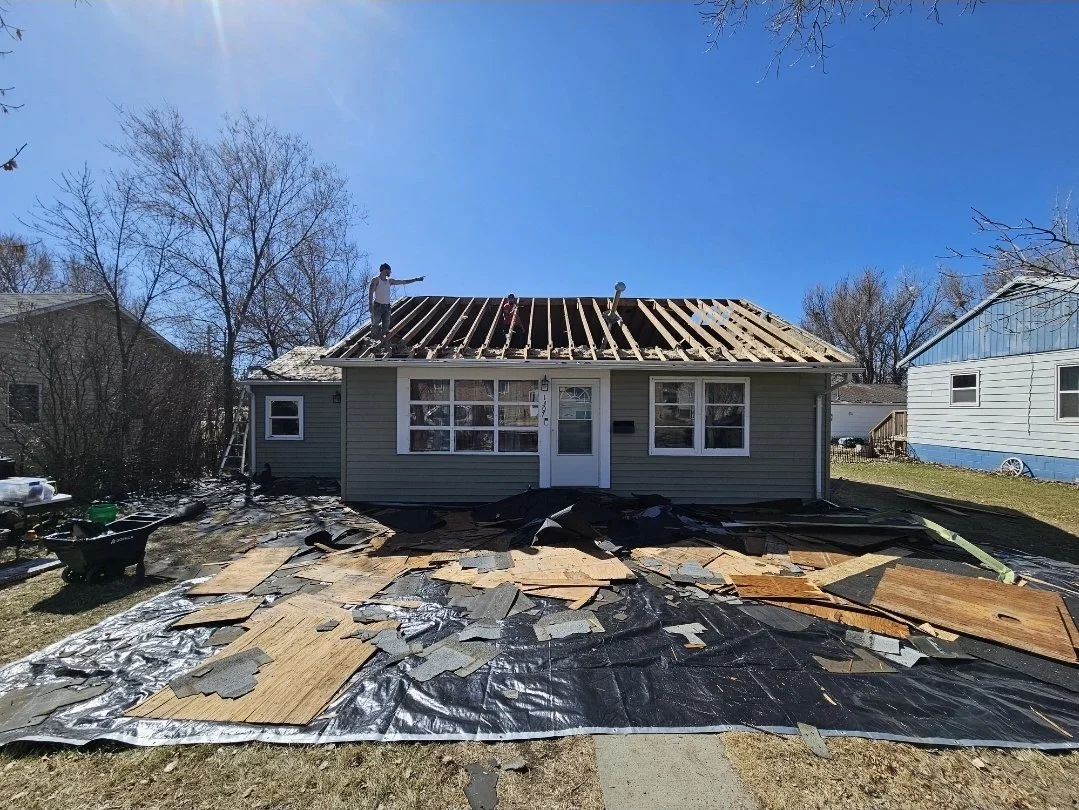 A house undergoing roof renovation with workers removing old shingles and framing the roof, surrounded by construction materials and tools.