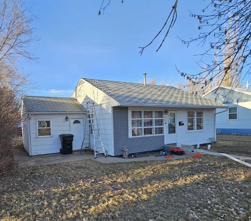 A house with a white exterior and a gray brick accent, partially painted, with a ladder leaning against it, construction tools on the ground, and a small backyard area with bare earth and a sidewalk.