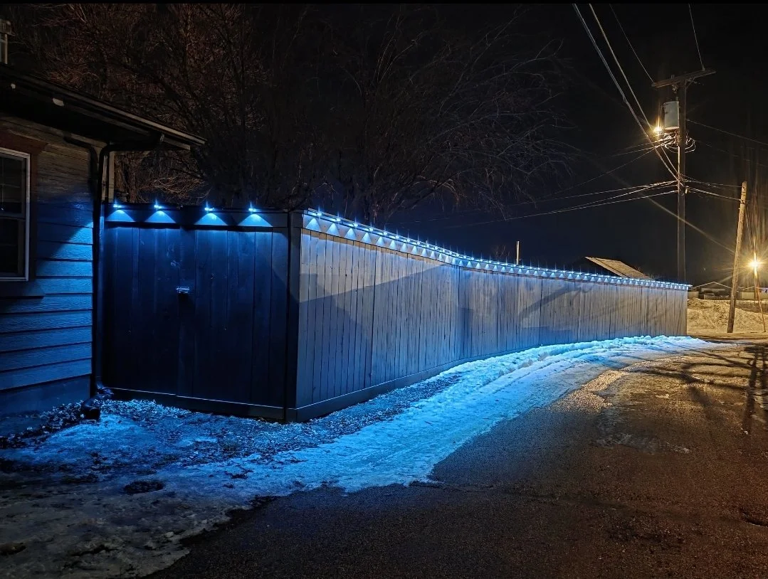Nighttime scene of a wooden fence decorated with blue LED lights along a street, with patches of snow on the ground and a house on the left side.