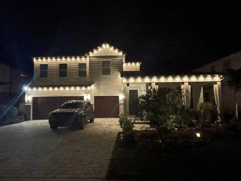 A two-story house decorated with string lights at night, with a driveway, parked SUV, and a front garden.