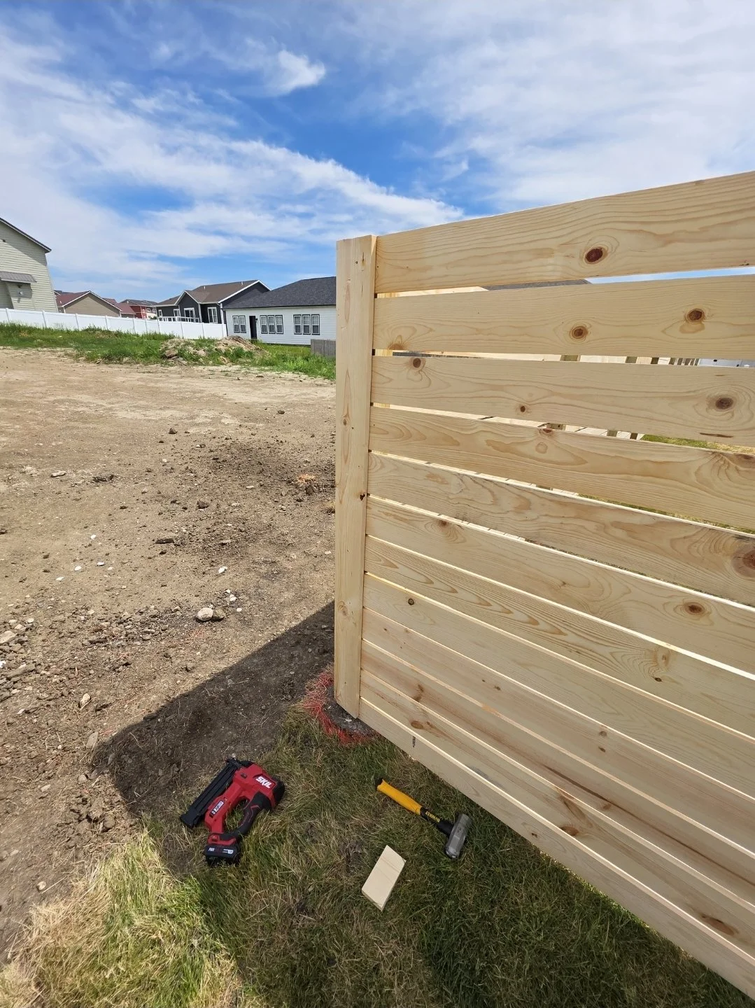 Construction site with a newly built wooden fence, tools on the ground including a drill and hammer, dirt ground, neighboring houses, under a partly cloudy sky.