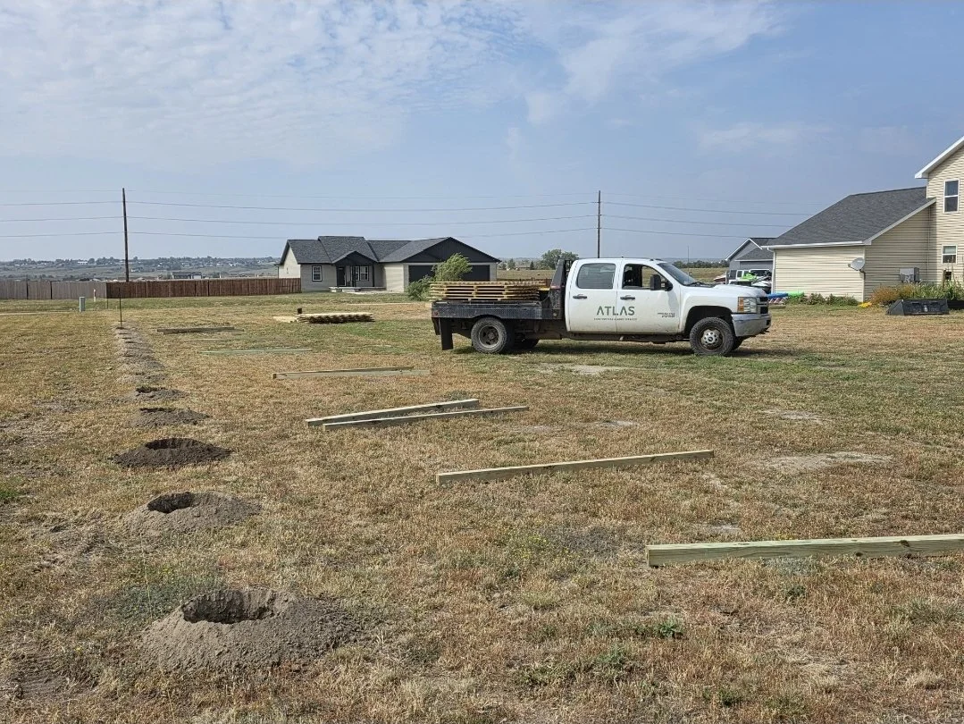 Construction site with dug holes and wooden planks, white pickup truck with company logo, in a suburban area with houses and open land.