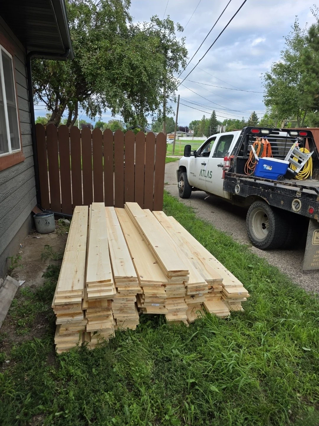 Stack of unfinished wooden boards or planks outdoors near a house, with a brown wooden fence, a pickup truck with tools in the bed, and trees and power lines in the background.