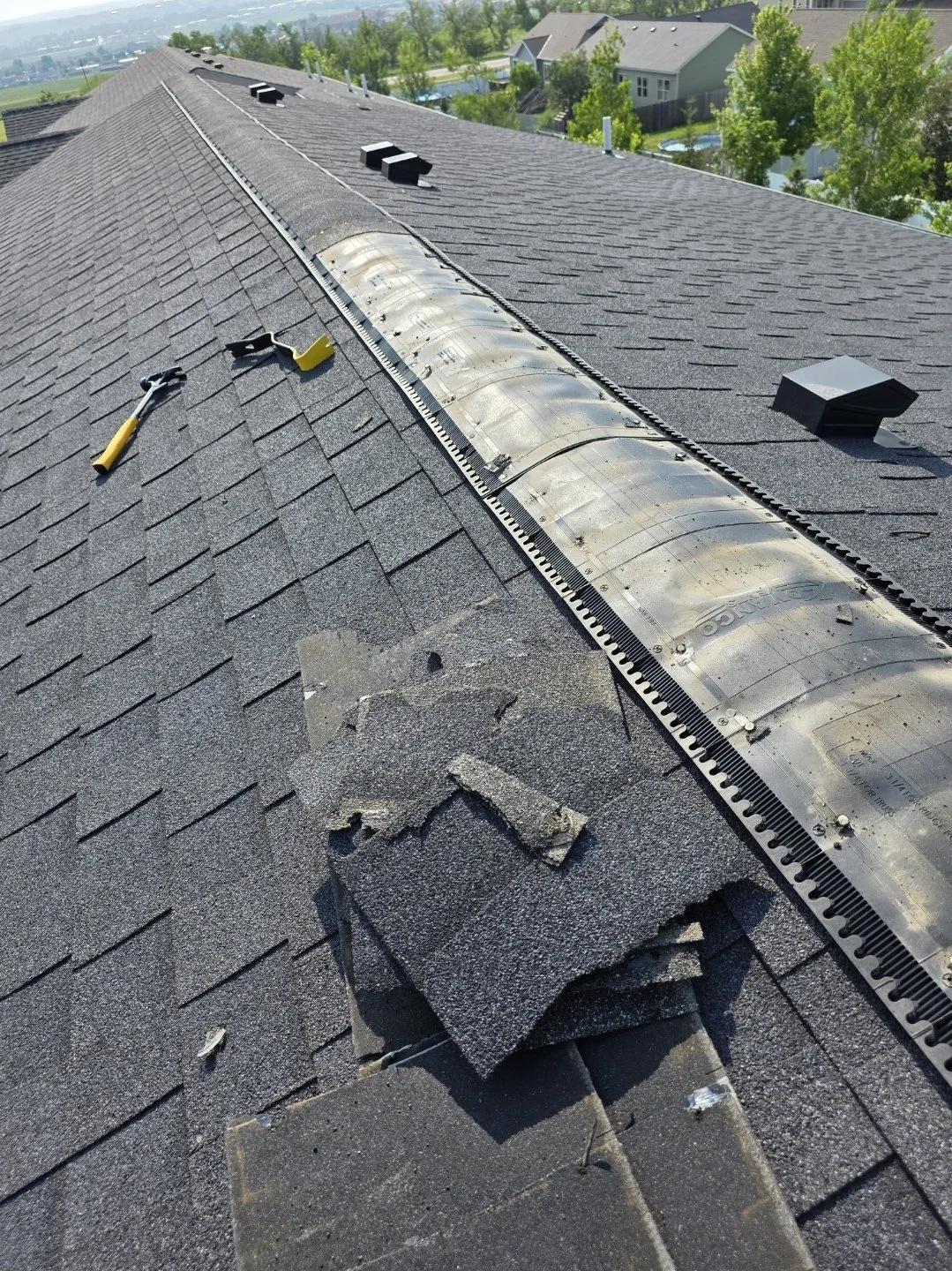 Close-up of a house roof undergoing maintenance, with shingles, tools, and a vent pipe visible, and in a suburban neighborhood with trees and other houses in the background.