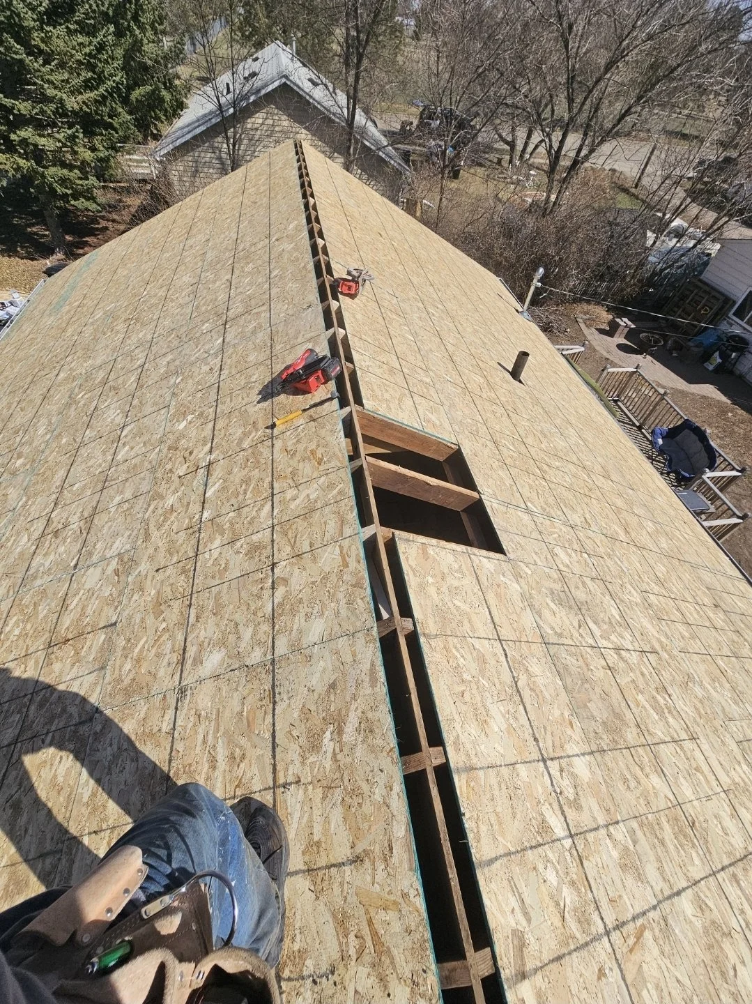Aerial view of a house roof under construction with plywood sheathing, two red power tools, and a worker's foot visible at the bottom of the image.
