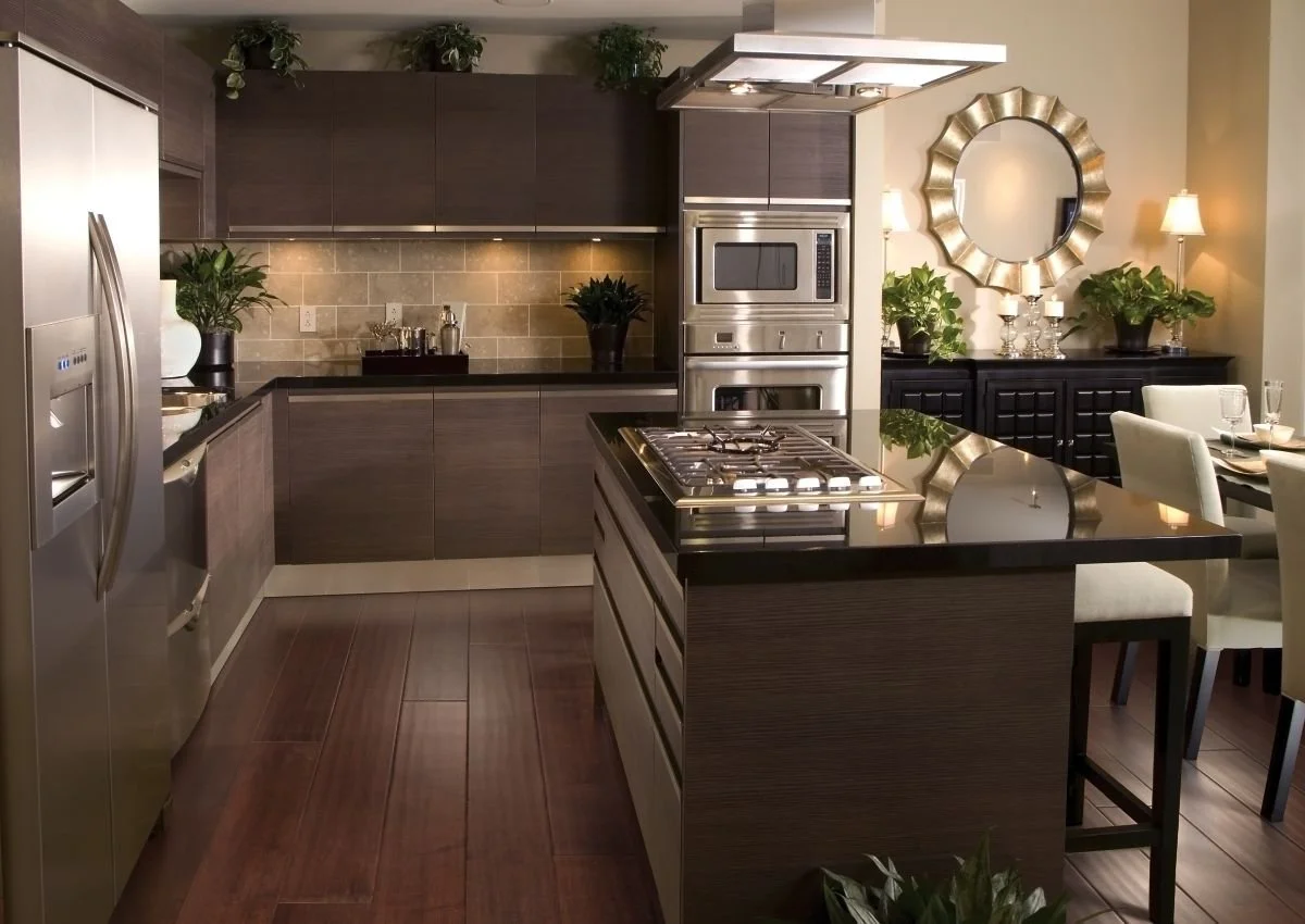 Modern kitchen with dark wood cabinets, stainless steel appliances, a gas stove on an island with black countertop, and a dining area with light-colored chairs and table. Decor includes potted plants, a large mirror, and wall-mounted lamps.