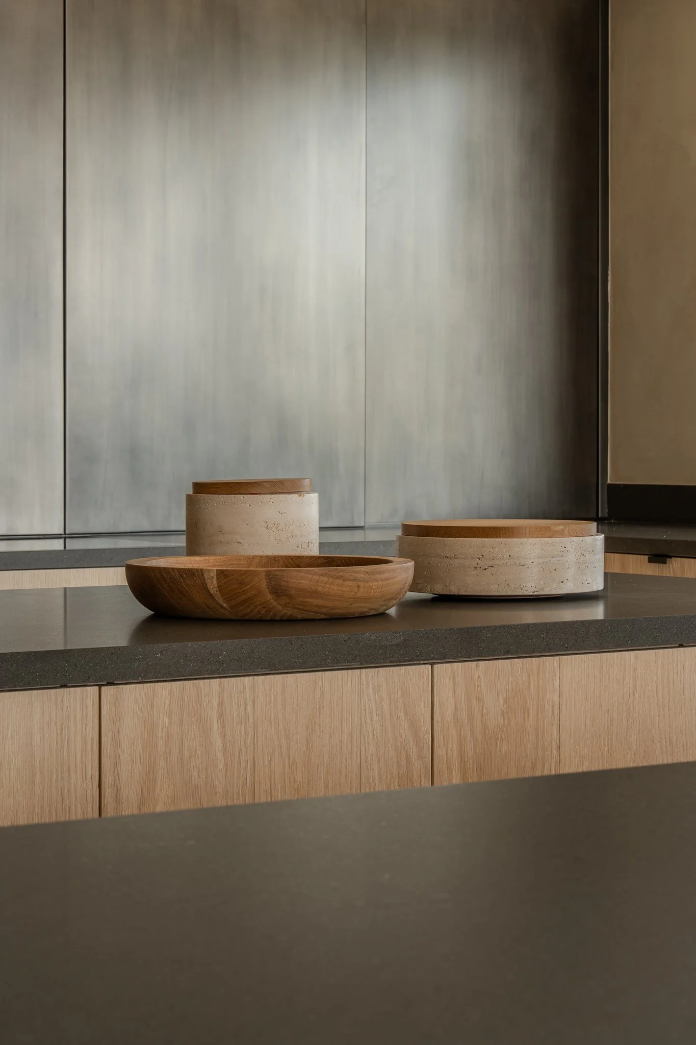 Wooden and concrete bowls and containers on a kitchen counter with wooden cabinetry and a metallic backsplash.
