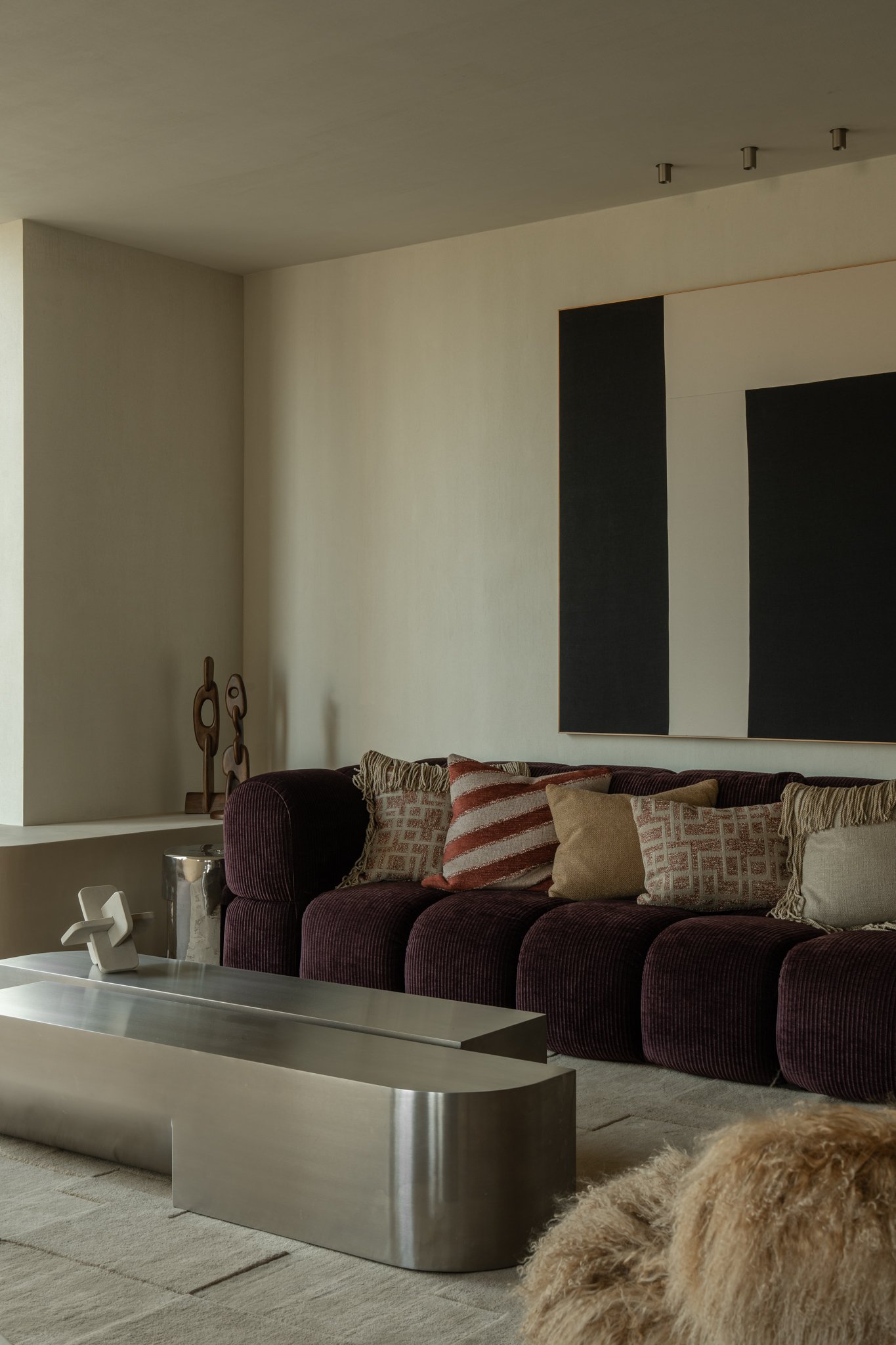 Modern living room with a dark purple velvet sofa adorned with various patterned throw pillows and a minimalist silver coffee table in front. There is a large abstract black and white artwork on the wall and a beige textured rug on the floor.