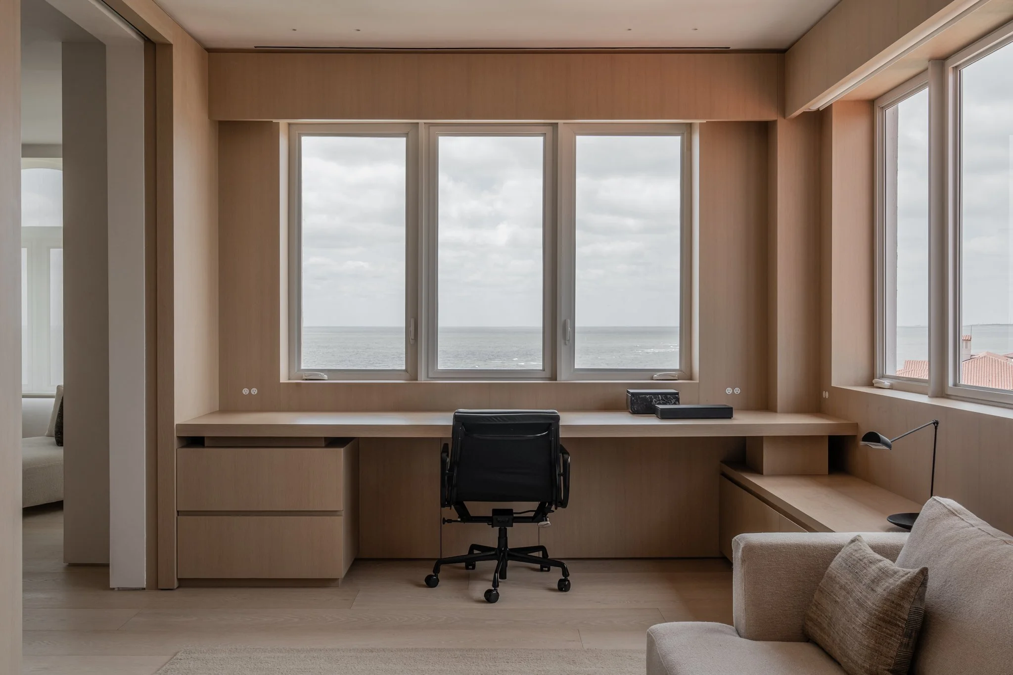A minimalist home office with a wooden desk under large windows overlooking the ocean, an black office chair, and a beige armchair with a cushion.