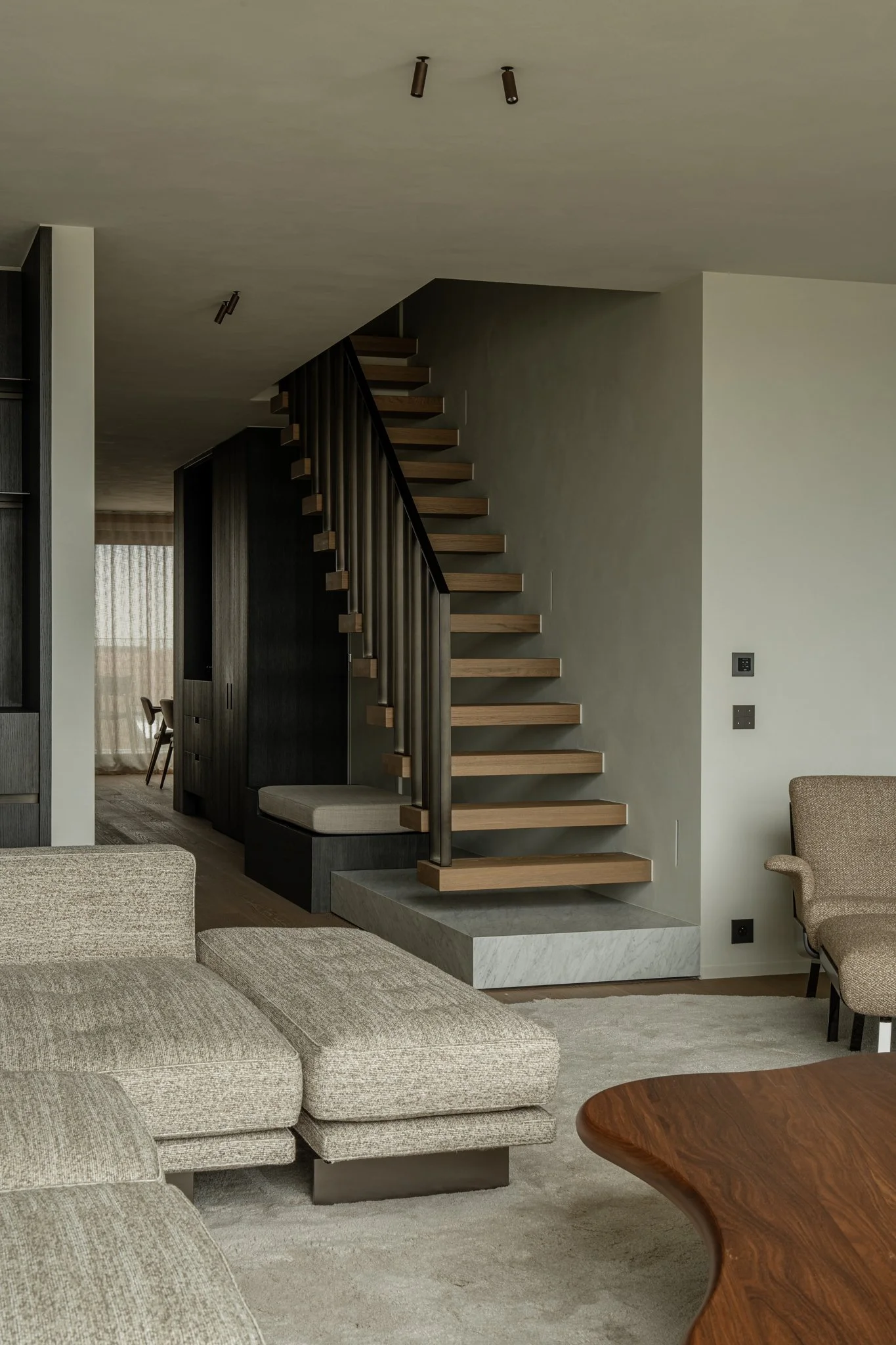 Modern living room with beige sectional sofa, wooden coffee table, and a floating staircase with wooden steps and black railing.