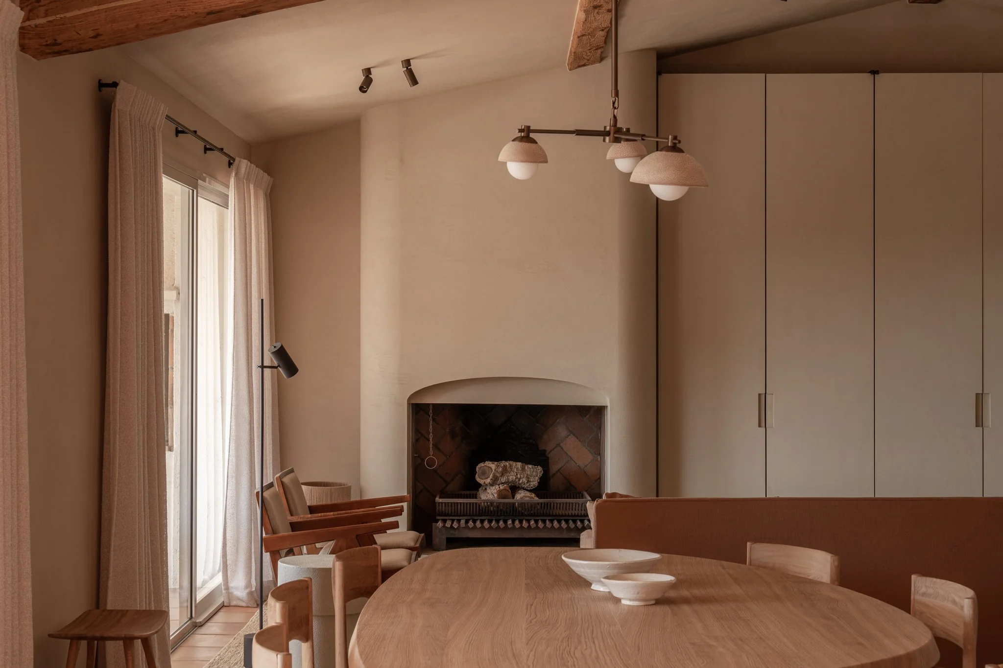 Interior view of a dining room with an oval wooden table, chairs, a fireplace with logs, beige curtains on a sliding glass door, and minimalist ceiling lighting.
