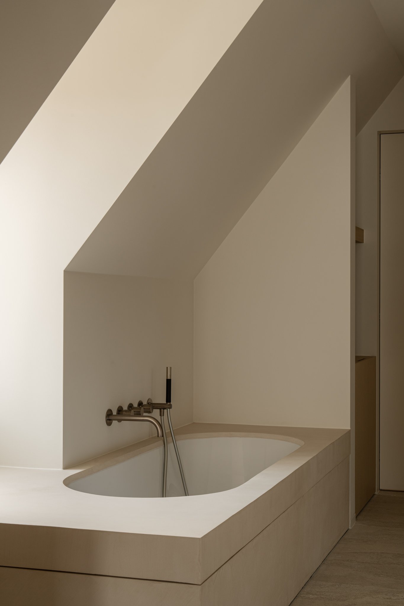 A minimalist bathroom with a built-in bathtub under sloped ceilings, with beige walls and floor, and a sleek wall-mounted faucet.