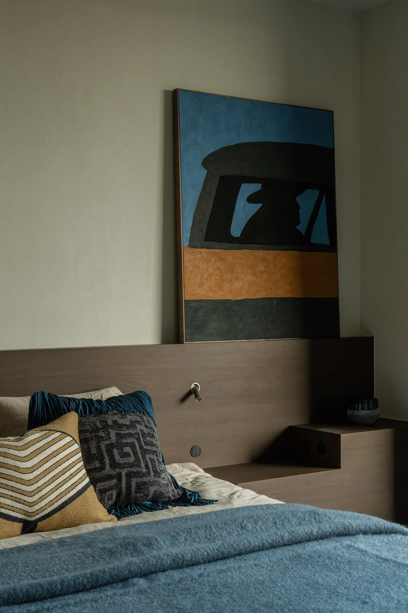 A bedroom with pillows on a bed, a wooden headboard with built-in side table, a bowl on the side table, and a large abstract painting hanging on the wall above the headboard.