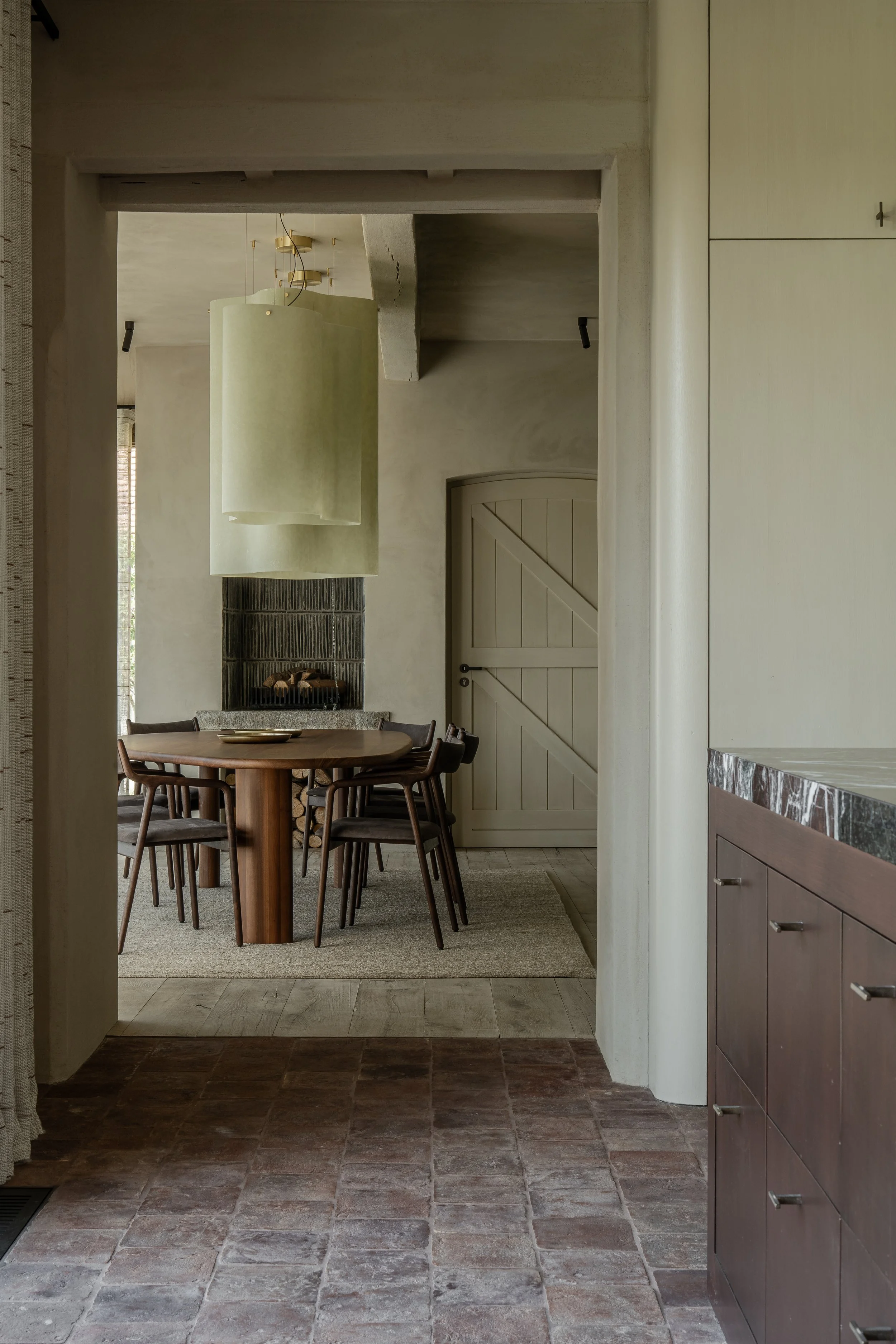 A view through an open door into a dining area with a round wooden table and six chairs, a fireplace with stacked firewood, a large hanging light fixture, and a wooden door with diagonal planks.