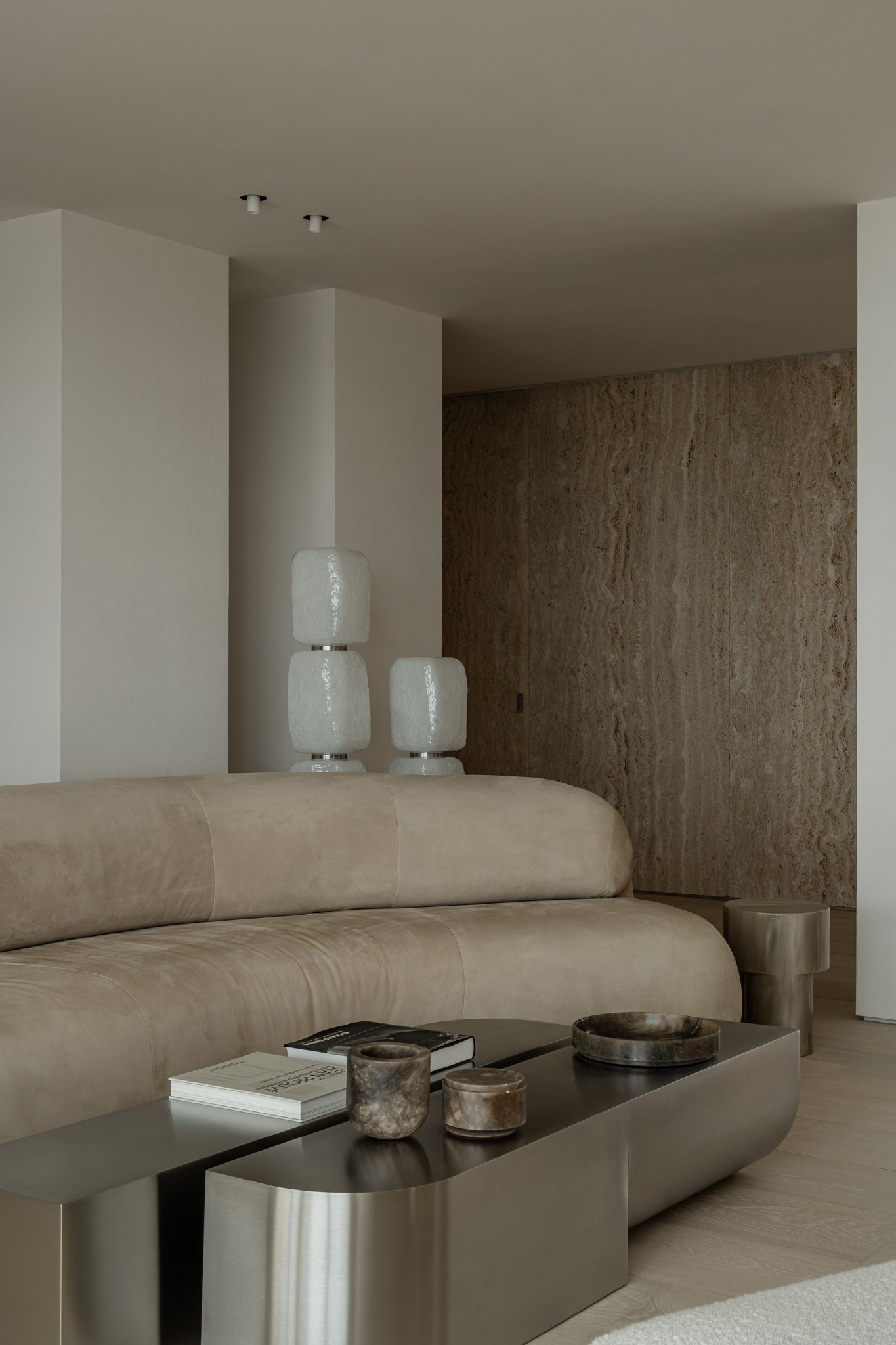 Minimalist living room with a beige curved sofa, black coffee table holding books and decorative objects, textured wall, and modern white lamps.