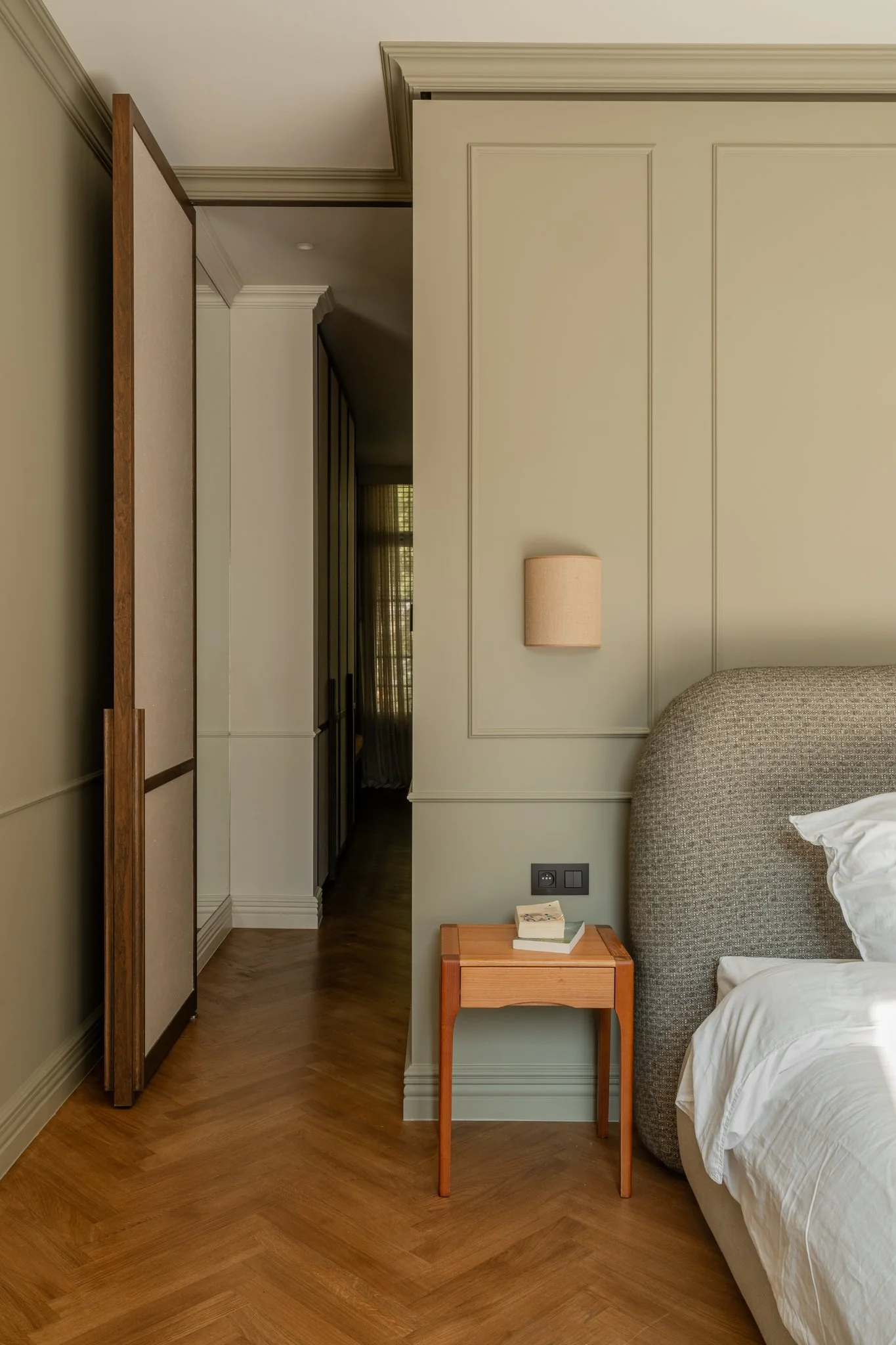 A bedroom with a nightstand next to a bed with a cushioned headboard and a wall-mounted sconce light. A hallway with dark wood flooring and a wood-paneled divider is visible in the background.