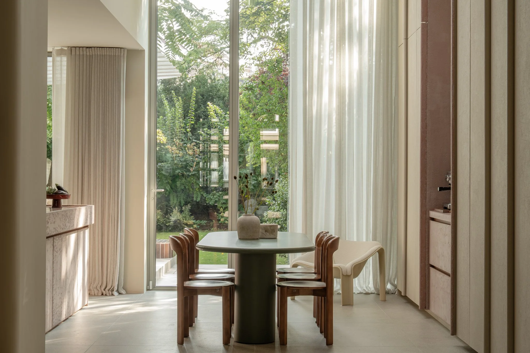 Bright dining area with a round table and six chairs, large glass window with sheer curtains, greenery outside, minimalist decor, and a modern aesthetic.