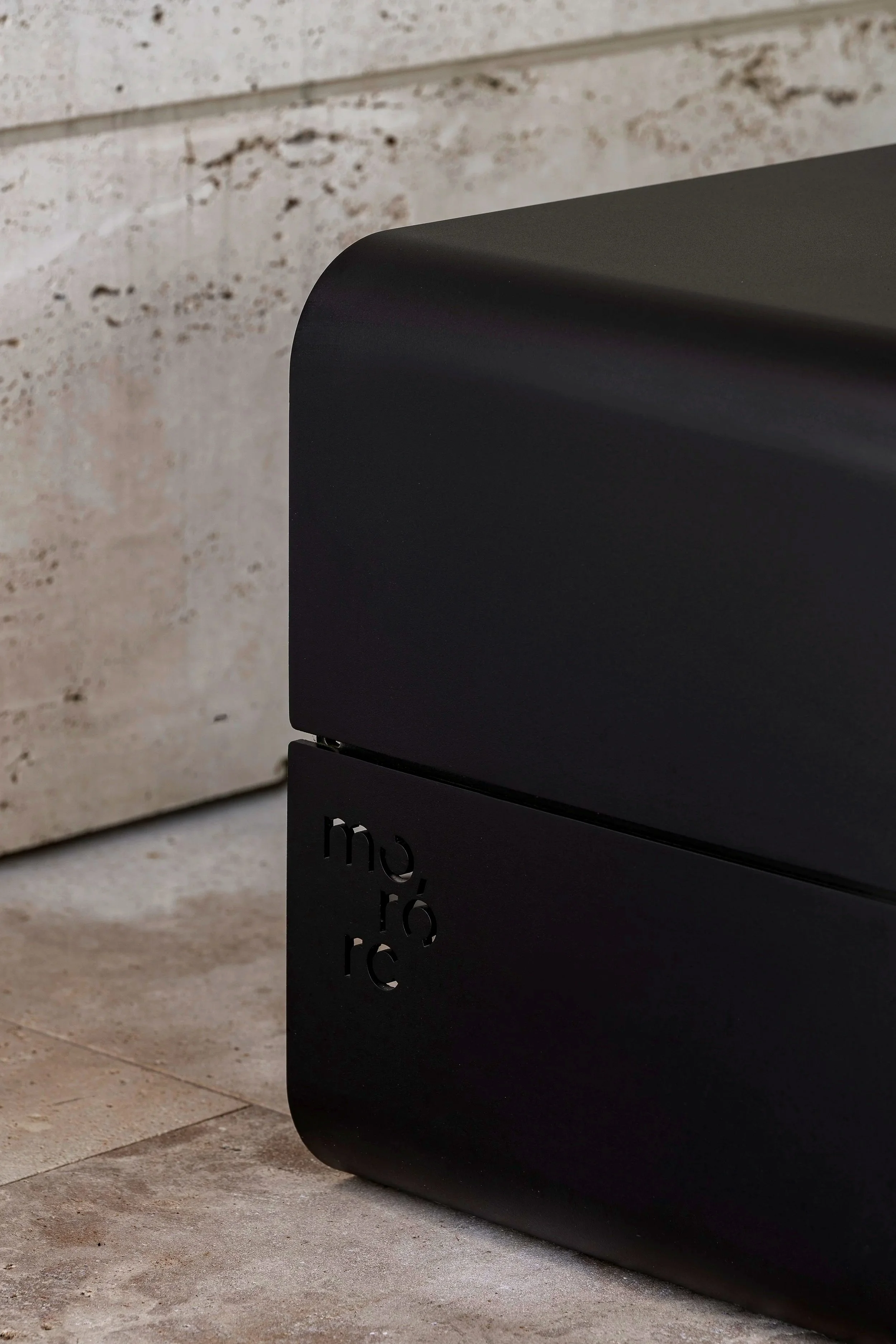 Close-up of a black modern speaker with a logo on the side, resting on a tiled floor near a textured wall.