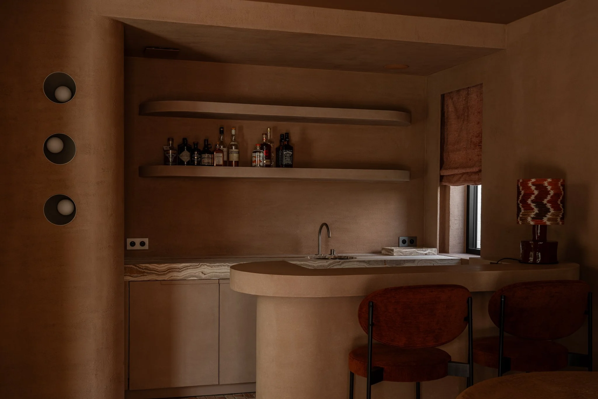 A small kitchen with wooden walls, two open shelves with bottles, a countertop with a sink, and a window with a brown Roman shade. There is a table with two orange chairs and a table lamp with a patterned shade.