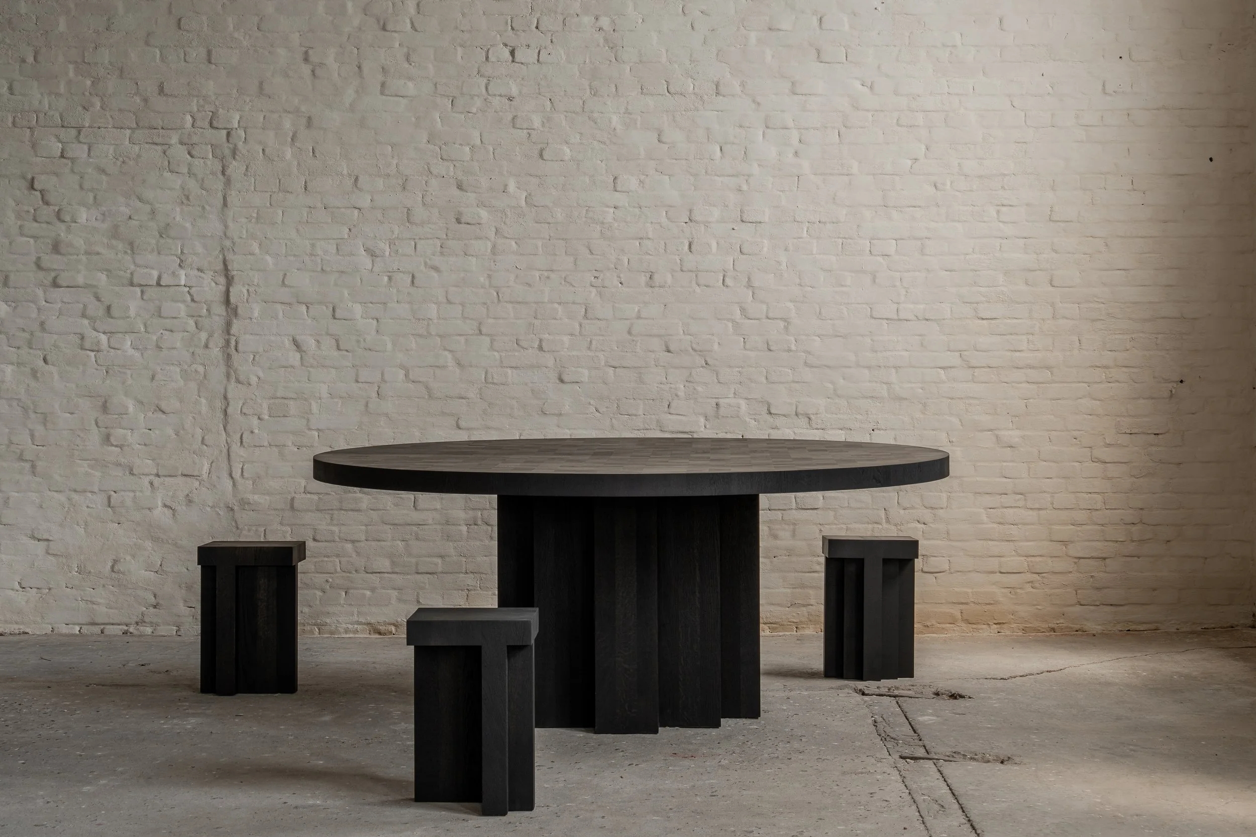 A round black wooden table with three matching black wooden stools in an industrial-style room with a white brick wall and concrete floor.